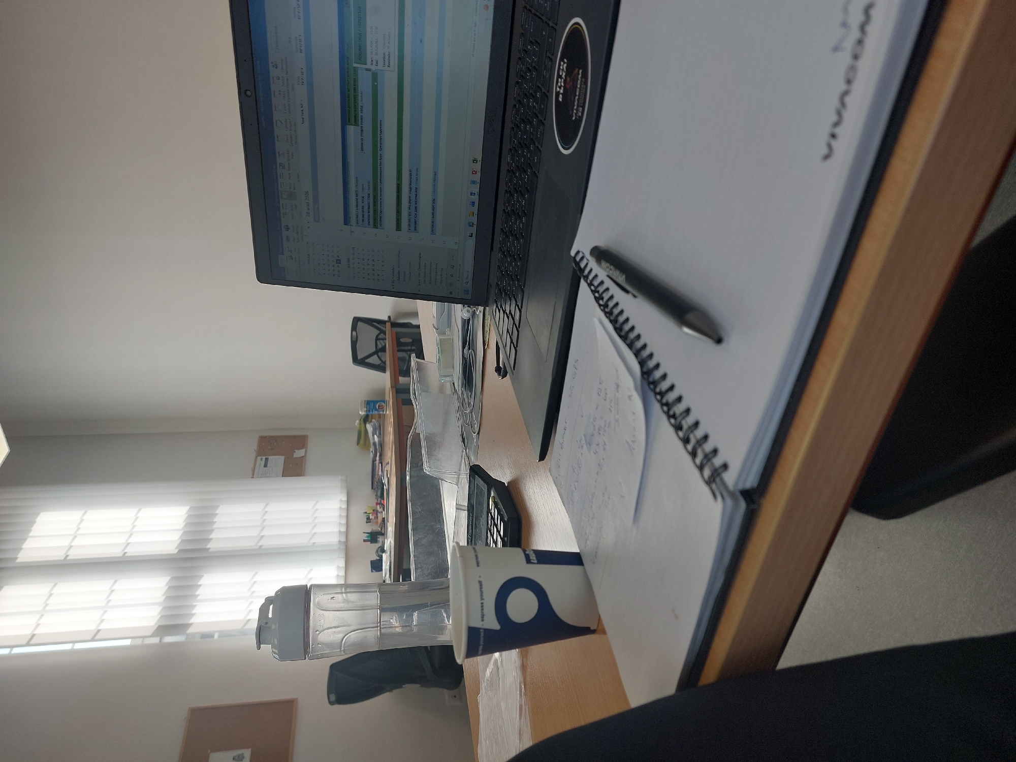 The image shows a desk setup. On the desk is a laptop displaying a calendar or schedule application. Beside the laptop, there's a notepad with the word "VIVACOM" visible on the cover. A pen rests on the notepad. A calculator and a water bottle are positioned near the laptop and notepad. A partially visible cup stands on the desk as well. The background features a window with blinds, with additional office supplies and what appears to be a corkboard on the wall. The scene appears to be a workspace, potentially an office. The OCR text indicates date information, possible location details ("New York, NT"), and text in Cyrillic script, suggesting a location in Bulgaria. Given "Сунгурларе Сдружение" in the OCR text and the prompt's request for Burgas, Bulgaria, the location might be within the Burgas Province, as Sungurlare is a town in that province.