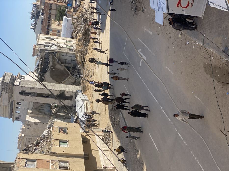 An aerial-perspective photograph captures a devastated urban street scene in Khan Yunis, Palestinian Territory, under a clear blue sky. A large, multi-story building, possibly a mosque or significant public structure, dominates the center, exhibiting severe structural compromise with a partially collapsed facade, extensive debris at its base, and significant damage to its upper sections, including a compromised domed or cupola element. To its immediate left, a multi-story residential or commercial building also displays significant damage, with debris scattered on its roof and within broken window frames. Other damaged structures, including one with apparent fire damage, are visible in the background.
The paved street running through the foreground and middle ground features faint white lane markings and a prominent light-colored hose or cable stretched across it. Over fifty individuals, comprising adults and children, are dispersed throughout the scene. They are primarily walking on the street and adjacent dirt paths, individually or in small groups. In the foreground, a man in a light-colored shirt walks, pulling the long hose. Piles of concrete, rebar, and other construction debris are abundant on both sides of the street, particularly around the bases of the damaged buildings. Several white tarpaulin shelters are erected on the dirt and rubble, with one in the lower right displaying red Arabic script. Dark utility lines traverse the upper portion of the frame.