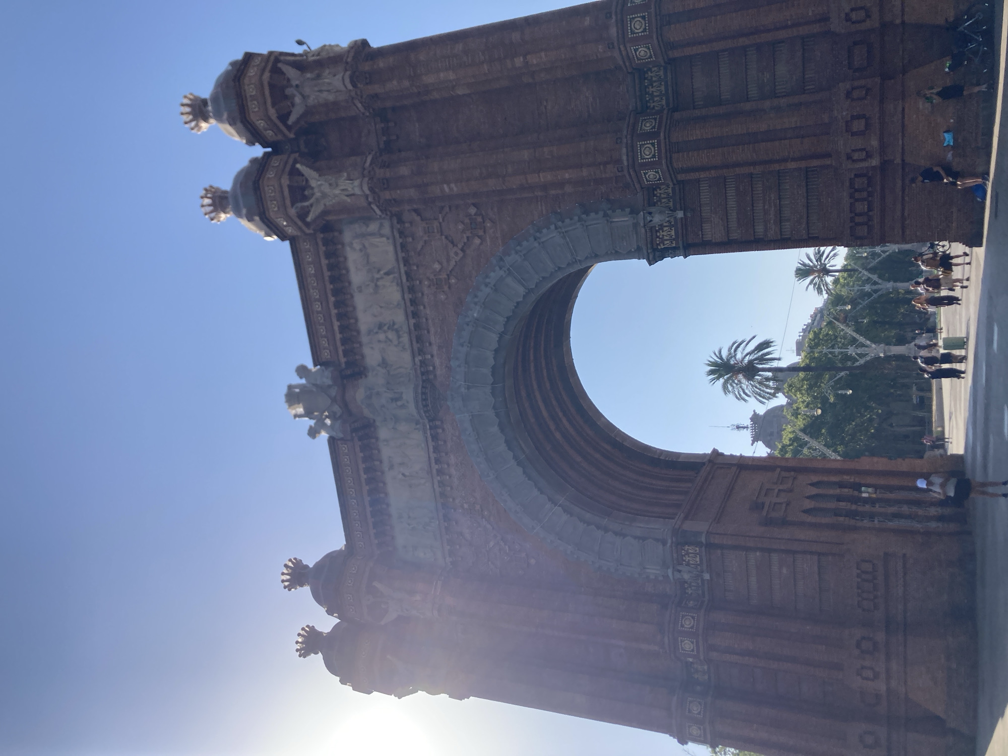The Arc de Triomf in Barcelona, Spain, is depicted in this low-angle, full-shot photograph taken on a clear, sunny day. The monumental arch, constructed of red brick and adorned with intricate carvings and sculptures, dominates the frame. Sunlight flares from the upper left, casting a bright glow on the left side of the arch and creating lens flare.
Beneath the arch, a palm tree is visible against a bright blue sky. In the background, a dense cluster of trees forms a green backdrop, with glimpses of buildings peeking through. Further into the distance, another palm tree stands tall.
The foreground shows a paved area with several individuals walking and standing. A person wearing a white cap and a dark skirt is standing near the base of the arch on the left. To the right, a person leans against the arch, and further to the right, a stroller is parked. A group of people are walking away from the arch in the middle ground. The overall atmosphere suggests a bustling tourist destination.
