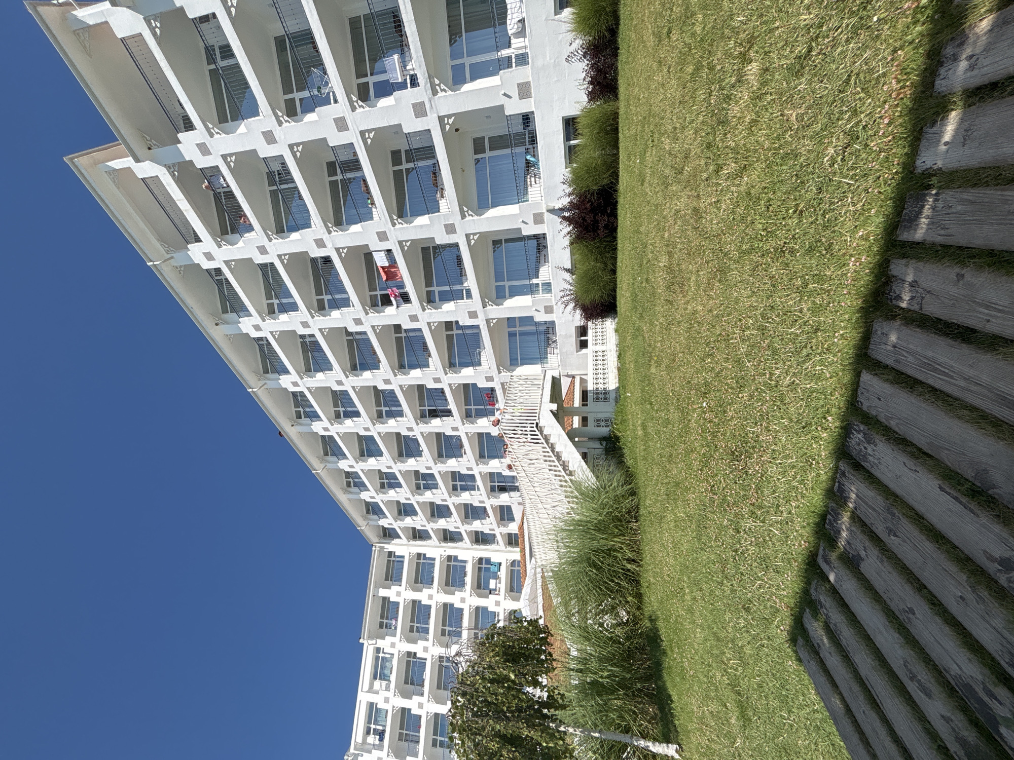 The image shows a white, multi-story hotel building with a curved facade, situated in Byala, Bulgaria. The building has numerous balconies with dark railings and large windows. The sky above is clear and blue. In the foreground, there is a well-maintained green lawn, with a pathway made of wooden planks in the lower left corner, casting a shadow on the grass. To the left of the building, there is a small tree with sparse foliage and some tall green grass. A white staircase with railings leads up to the hotel entrance. Some laundry is visible on several balconies. A few people are visible on the stairs and balconies. The overall scene suggests a bright, sunny day at a coastal resort.