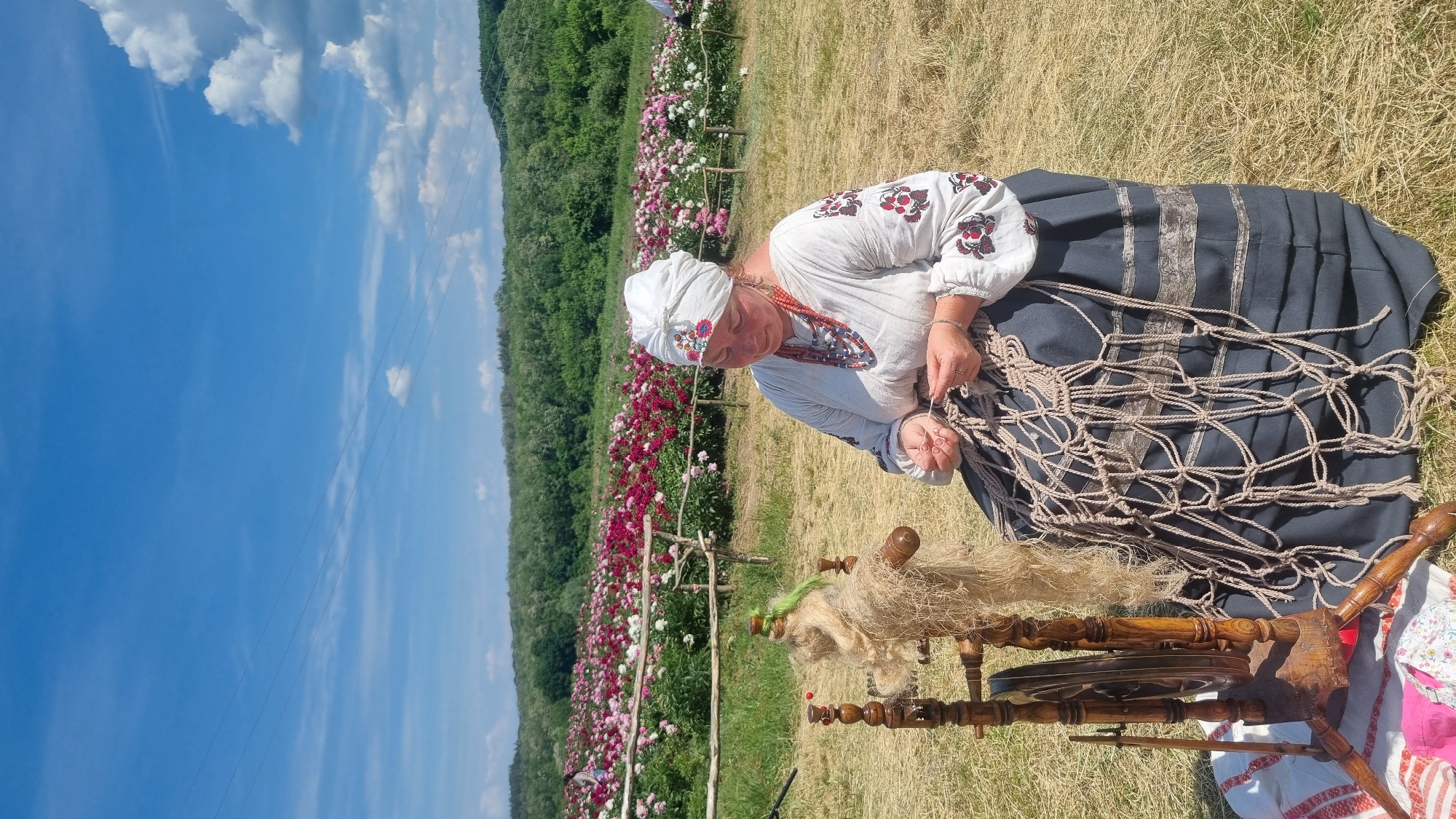 An outdoor scene in Melnyky, Ukraine, features a woman in traditional attire engaged in a craft on a sunny day. The woman, appearing middle-aged, is seated in the foreground, facing slightly right, with her gaze directed towards her hands. She wears a white headscarf with floral embroidery, a white long-sleeved shirt (vyshyvanka) with red and dark embroidery on the sleeves, a red beaded necklace, and additional multi-colored beaded necklaces. Her dark-colored skirt has lighter horizontal bands. She is actively working on a net-like structure made of light-colored twine, using her hands to create knots. To her immediate left, a traditional wooden spinning wheel holds a mass of light-brown, natural fiber, possibly flax or hemp, with a small green element intertwined. A red and white patterned cloth is partially visible on the ground beneath the spinning wheel. The ground consists of dry, yellowed grass. In the midground, beyond a rustic wooden fence, lies a large field of blooming peonies in various shades of pink, red, and white. The background features a dense line of green trees and foliage on a gentle slope. The sky above is clear blue with scattered white cumulus clouds, particularly dense on the right side. Power lines are visible stretching across the upper left portion of the sky.