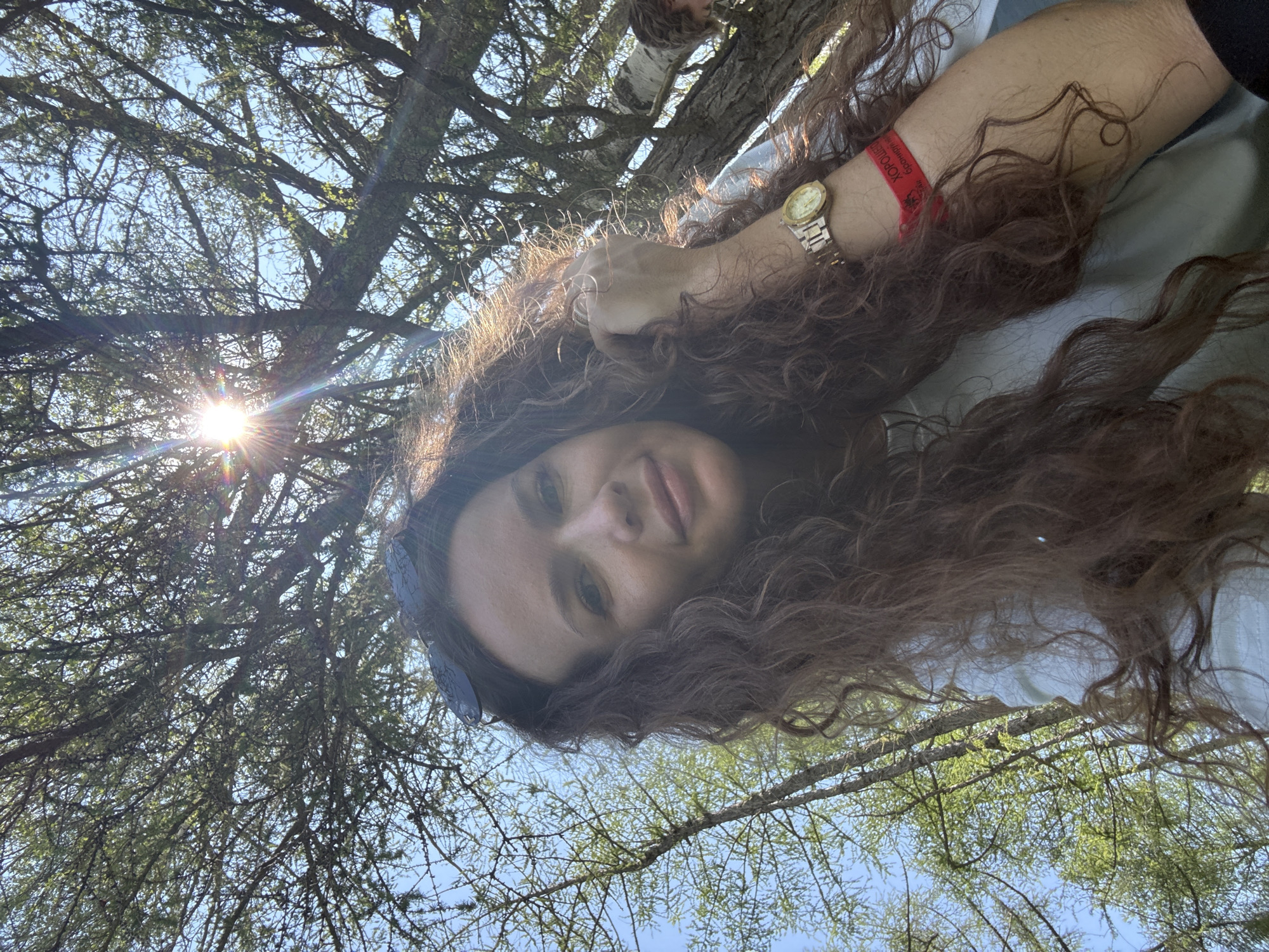 A low-angle, medium shot captures a woman with long, curly brown hair and sunglasses perched on her head, looking up towards the sun filtered through the branches of a larch tree. She is wearing a light-colored t-shirt and a red wristband with Cyrillic text that appears to read "Хорошо Быть" (It's Good to Be). A gold watch is on her left wrist. The background consists of a bright blue sky and numerous branches and needles of the larch tree. Another person, possibly a child, is partially visible in the upper right background, sitting in the tree. The scene appears to be outdoors, in a wooded area, potentially at the Bichiktu-Boom location in Russia, given the context.