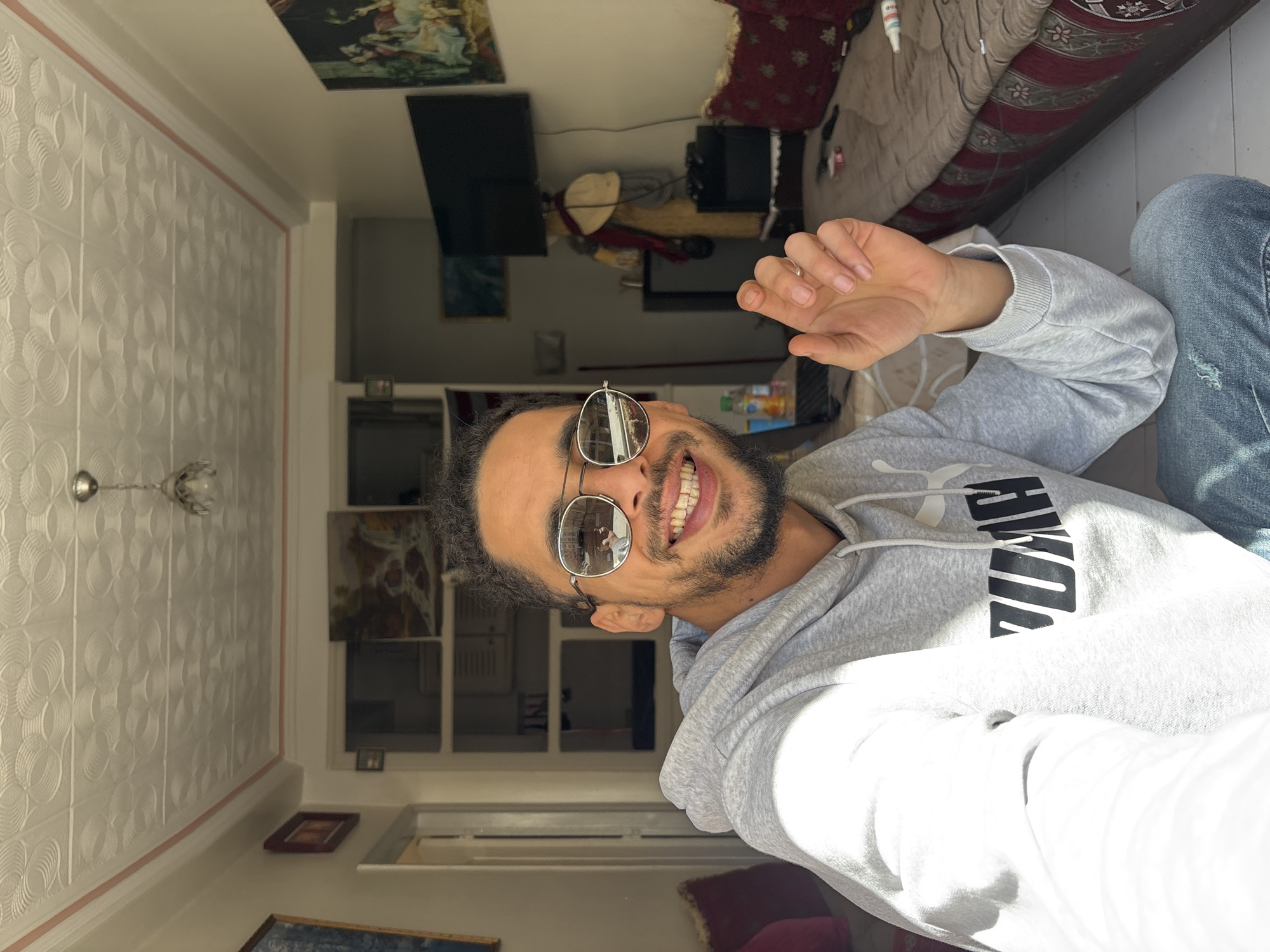 The image displays a male individual in a selfie-style shot within a residential interior, specifically located in Tanger, Morocco. The man, positioned in the mid-foreground, has dark, curly hair and a beard. He is wearing mirrored aviator-style sunglasses, a light grey hooded sweatshirt with a black "PUMA" logo on the chest, and distressed blue jeans. He smiles broadly at the camera, revealing his teeth. His right hand is raised, with fingers slightly curled, and a silver ring is visible on his right ring finger. The reflection in his sunglasses shows his left arm holding a device, indicative of taking the photograph.
The background features white walls and a white ceiling adorned with a repeating circular embossed pattern, bordered by a thin pink strip. A small, simple light fixture hangs from the ceiling. To the man's left, a dark-framed picture is partially visible on the wall, and a doorway leads to another room. A red cushion, likely part of a sofa, is also visible.
To the man's right, a traditional Moroccan seating unit (sedari) with a reddish-brown fabric cover patterned with gold and white motifs extends along the wall. Red cushions are placed on the sedari. A flat-screen television is mounted on the wall above a small, dark table. Adjacent to the table, a standing decorative item, possibly a mannequin, is dressed in a light-colored hat and a red scarf. Various small objects, including a remote control and a white tube, rest on the sedari. Behind the man's arm, two bottles (one green, one clear) are visible on a surface. Further back, a window with a white frame is visible, alongside two framed paintings: one depicting a landscape with a waterfall, and another showcasing a classical scene with multiple figures. Bright sunlight enters the room from the left, casting distinct highlights and shadows. The floor is tiled with white squares.