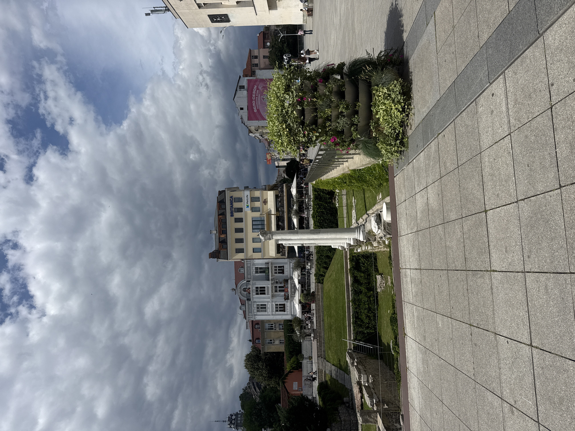 The image shows the Roman Stadium of Plovdiv, Bulgaria, on a cloudy day. In the foreground, a paved walkway is visible, leading towards a large tiered planter filled with greenery. Beyond the planter, the ruins of the Roman Stadium are apparent, with sections of stone walls and grassy areas. In the mid-ground, several white columns stand, remnants of the ancient structure. Behind the stadium, a row of buildings line a street, some with outdoor seating and umbrellas. The buildings are of varying architectural styles and colors, with one notable building featuring a sign for "Britannica" and "MetLife." In the background, more buildings and trees are visible, as well as a tall tower on a hill to the left. People are scattered throughout the scene, walking on the paved areas and near the buildings. The sky is filled with dramatic, dark clouds, suggesting an impending storm. A pink banner with Cyrillic text and the year "2026" is displayed on one of the buildings.