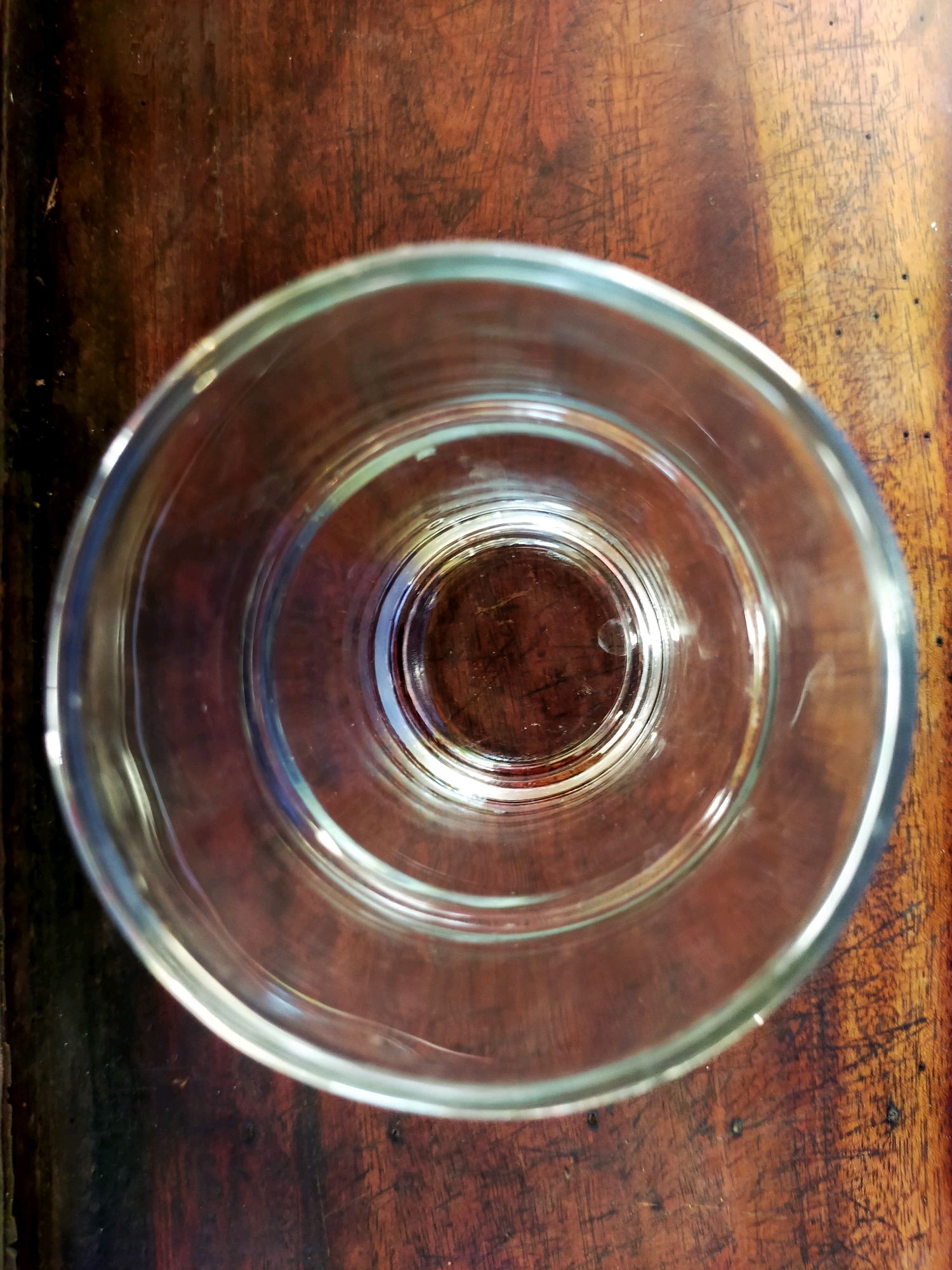 A top-down, medium shot captures a clear glass tumbler resting on a dark brown wooden surface. The tumbler is empty, and its interior reveals concentric rings, a design feature of the glass. The wooden surface appears worn, with visible grain and some small indentations or marks. The lighting creates a subtle reflection on the glass and highlights the texture of the wood. No people or other objects are visible in the frame. The scene is set in an unspecified indoor location, possibly a home or a cafe, in Surabaya, Indonesia.