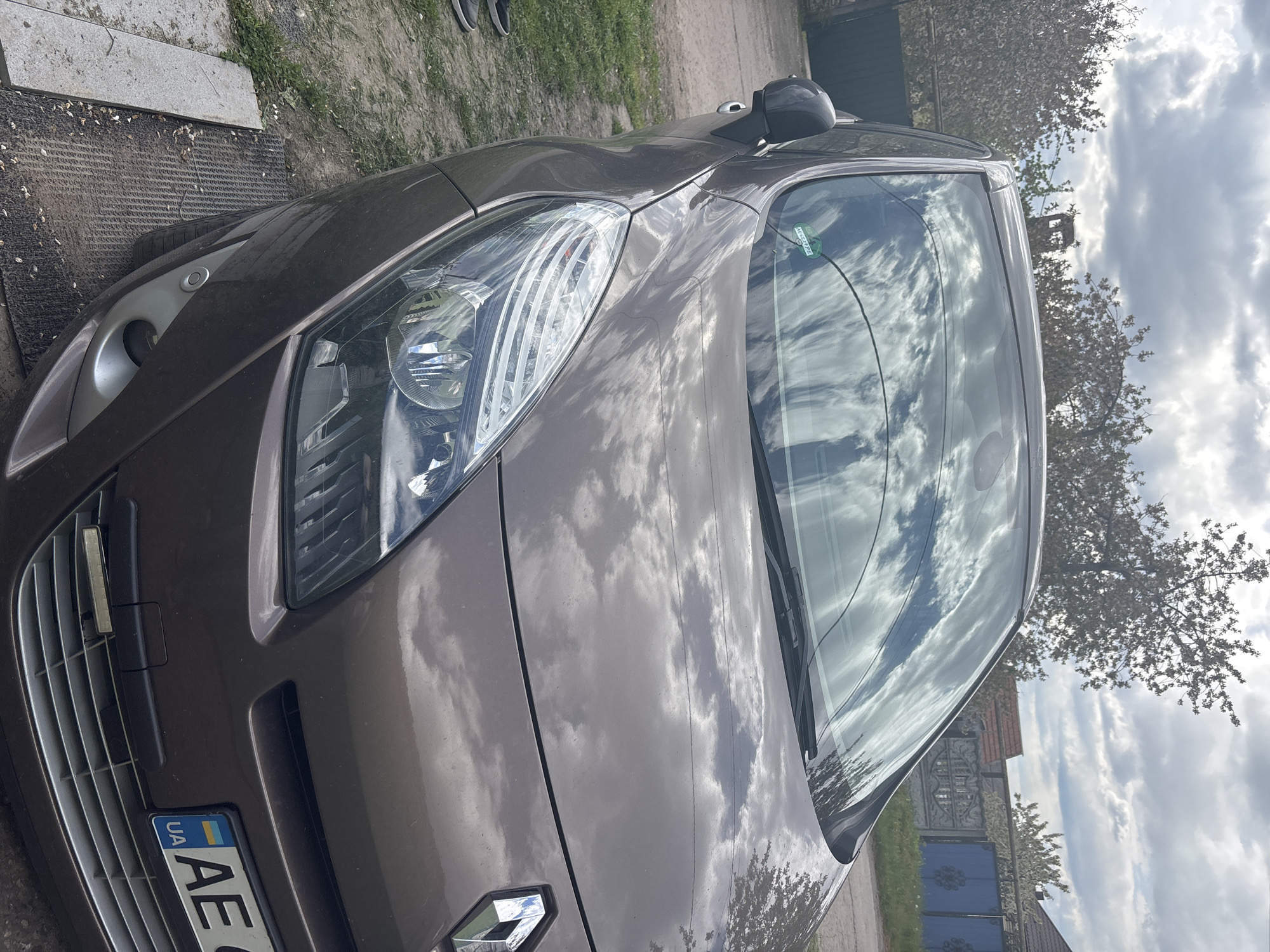 The image, rotated 90 degrees clockwise, displays the front and passenger side of a brown or bronze car, likely a Renault given the visible logo. The car is parked outdoors on a dirt and gravel surface, with concrete slabs and sparse grass nearby.
The car's body and windshield prominently reflect a cloudy sky and the surrounding environment, including trees, some of which appear to be flowering. Notable car features include clear headlights, a front grille, and a small, round sensor on the front bumper. A Ukrainian license plate is visible, showing "UA" and "AE". On the passenger side of the windshield, a green sticker with the text "AL 2322 FL" is affixed.
In the background, beyond the car, are more trees, a fence, and part of a building with a dark gate. The overall scene is brightly lit, indicating daytime under a heavily clouded sky. While no specific activity is taking place, the car appears to be stationary. A pair of feet in sneakers is partially visible near the front of the car, suggesting someone is standing close by. The setting appears to be a driveway or unpaved area, possibly in a suburban or rural location.