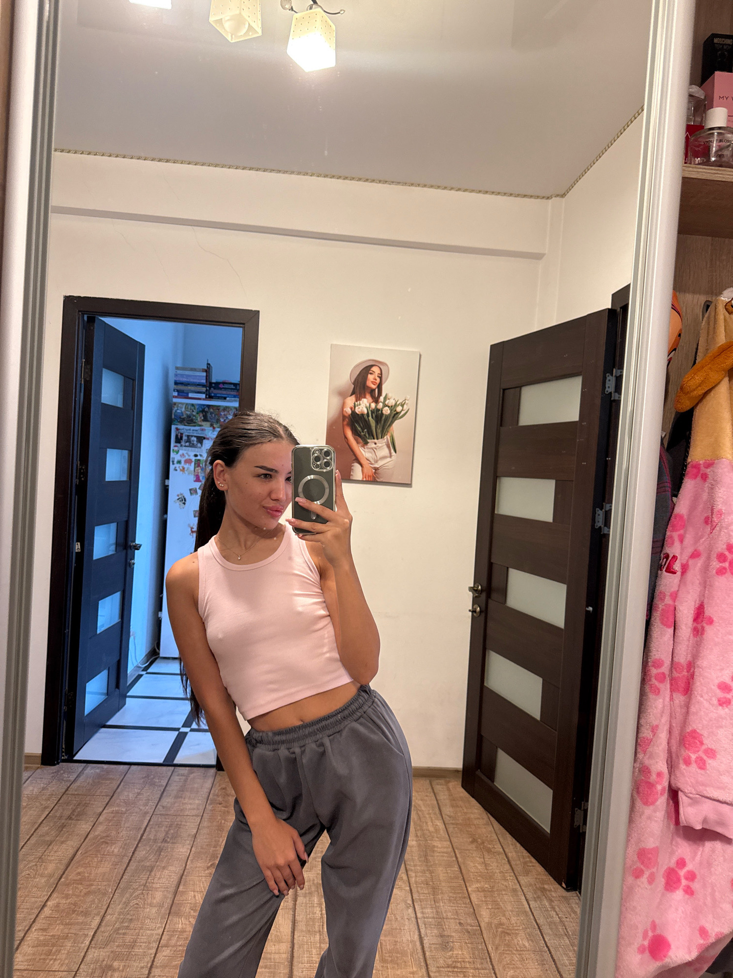 The image shows a young woman taking a selfie in a room with wood-paneled flooring and two dark wood doors. She is wearing a light pink tank top and grey sweatpants. In the background, there are several framed pictures on the wall and a closet with clothing. The room appears to be a domestic interior, possibly an apartment or house.