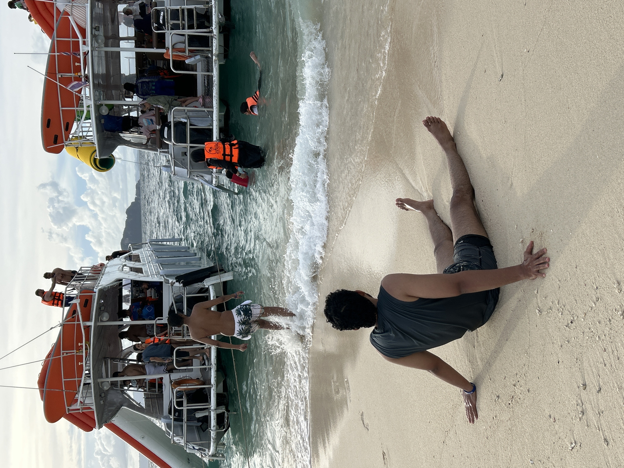 In Phru Nai, Thailand, on a sandy beach adjacent to clear, turquoise waters, a man is seated on the sand, facing away from the camera, with his legs extended forward. He is wearing a dark, sleeveless shirt and dark shorts. Gentle waves lap at the shore. In the background, two large, white tour boats with orange inflatable slides are anchored in the water. Several people are visible on and around the boats. One man, wearing swim trunks, is wading in the shallow water near the shore, with waves splashing around his legs. Another person in the water is wearing an orange life vest and appears to be swimming. People are also seen on the decks of the boats, some wearing orange life vests, and engaging in various activities, suggesting a recreational excursion. The sky is partly cloudy, with a soft, diffused light.