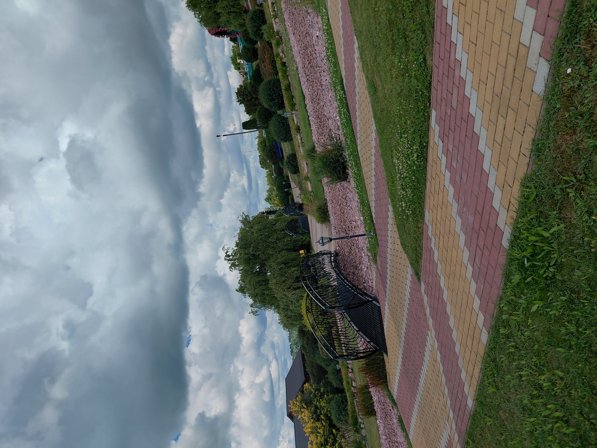 The image depicts an outdoor park scene under a cloudy sky. In the foreground, a pathway paved with red, yellow, and gray interlocking stones winds through lush green grass. To the left and center, a black ornamental metal bridge with intricate scrollwork spans across a dry streambed filled with light pink stones. A lamppost stands near the bridge. In the background, a large weeping willow tree dominates the landscape, with a building featuring a dark brown roof visible behind it. Further in the distance, more trees, bushes, and decorative plants are arranged on a tiered hillside. The scene appears to be a well-maintained public space, likely in the city of Kovalivka, Ukraine.