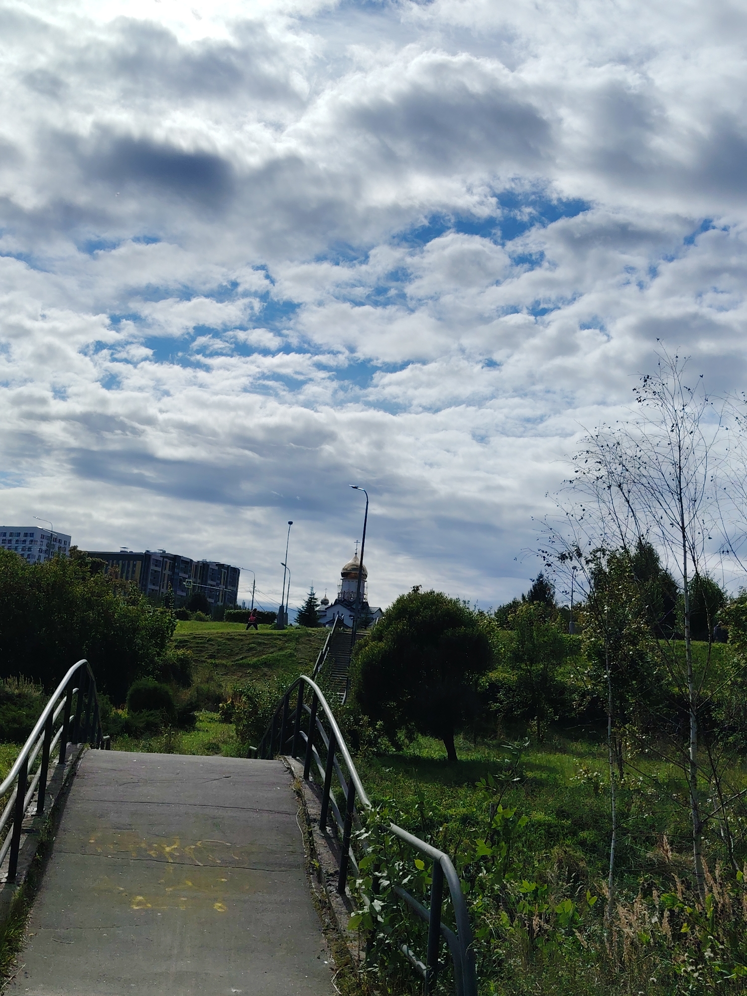 An overcast day with dramatic, puffy clouds is depicted. The image shows a park-like area with a concrete bridge leading uphill. Beyond the bridge, there is a grassy slope with trees and bushes. In the background, there are several multi-story buildings, a lamppost, and a golden-domed building that appears to be a church, possibly the Church of St. Sergius of Radonezh in Zelenograd, Russia, given the architectural style and its typical placement within green spaces in the area. A lone figure in red is visible walking up the grassy incline. The overall scene suggests a tranquil, possibly urban recreational space.