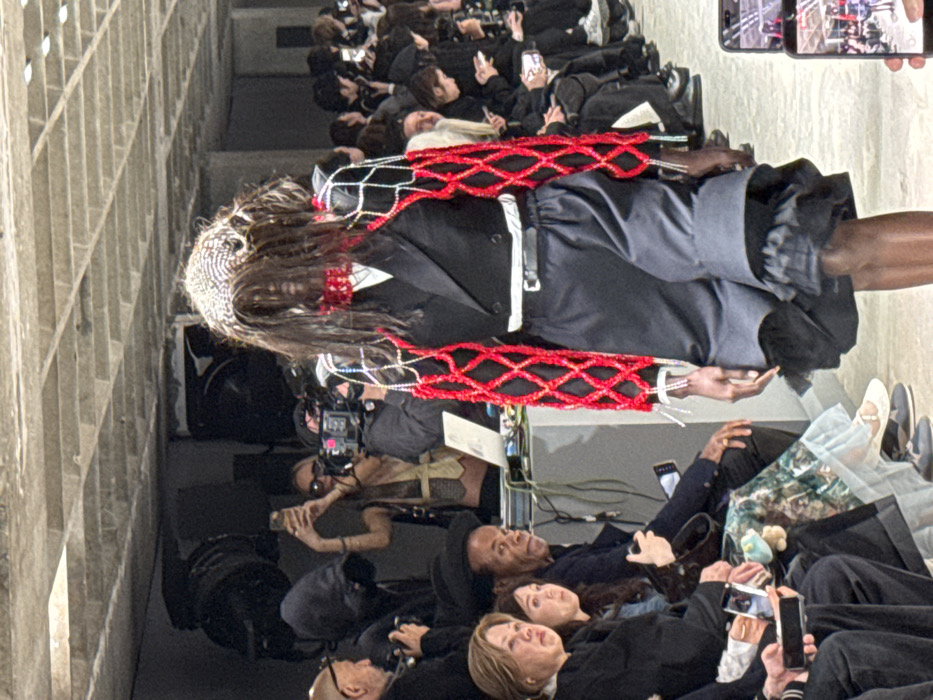 A fashion show is underway in a large, industrial-style venue with a concrete ribbed ceiling in Paris, France. The primary subject is a dark-skinned model walking left-to-right on a light-colored runway, directly facing the camera. The model wears a striking ensemble: a headpiece of intertwined dark hair and a beaded silver mesh, a red beaded choker, a black cropped double-breasted blazer, and an inner white band. Attached to the blazer are voluminous, long sleeves constructed from red, diamond-patterned netting heavily embellished with dangling white beads/pearls. Black, loose-fitting knee-length shorts with a ruffled dark layer beneath the hem are worn with a black belt.
Seated along the runway to the left are numerous audience members. An older woman with light brown hair and a younger woman with dark hair are prominently visible in the mid-ground, observing the model. Further back, a dark-skinned man with short hair, wearing a dark jacket, intently watches the runway. Many spectators hold smartphones to record or photograph the event. In the background on the left, a blonde-haired woman in a light vest records with a smartphone, while a professional videographer operates a large camera on a tripod. Technical equipment, including a laptop and wires, rests on a gray console near the seated audience. The environment is utilitarian, featuring industrial lighting fixtures mounted on the concrete ceiling. Additional audience members are visible on the right, also engaged with the show.