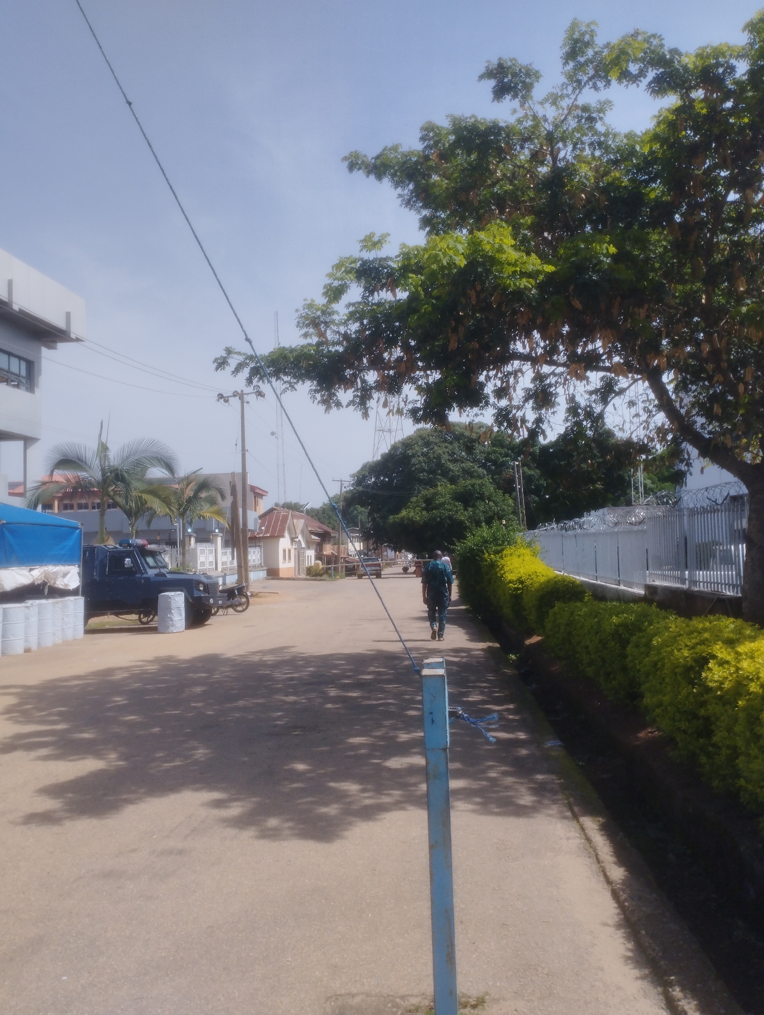 The image depicts a street scene in Jos, Nigeria, under a clear sky. In the foreground, a paved road leads into the distance, with tree shadows dappled across its surface. To the right, a neatly trimmed hedge of vibrant green and yellow bushes lines a pathway, bordered by a metal fence with barbed wire at the top. Further down the street, several low-rise buildings with tiled roofs are visible. On the left side of the road, a dark blue utility vehicle with a light bar on its roof is parked near a blue tarp-covered structure, possibly a truck bed, with white cylindrical objects stacked beneath it. A lone individual, dressed in dark clothing, walks away from the camera on the pathway to the right. Utility poles and wires are present throughout the scene, indicating an urban setting. The overall impression is that of a relatively quiet, well-maintained street within a Nigerian city.