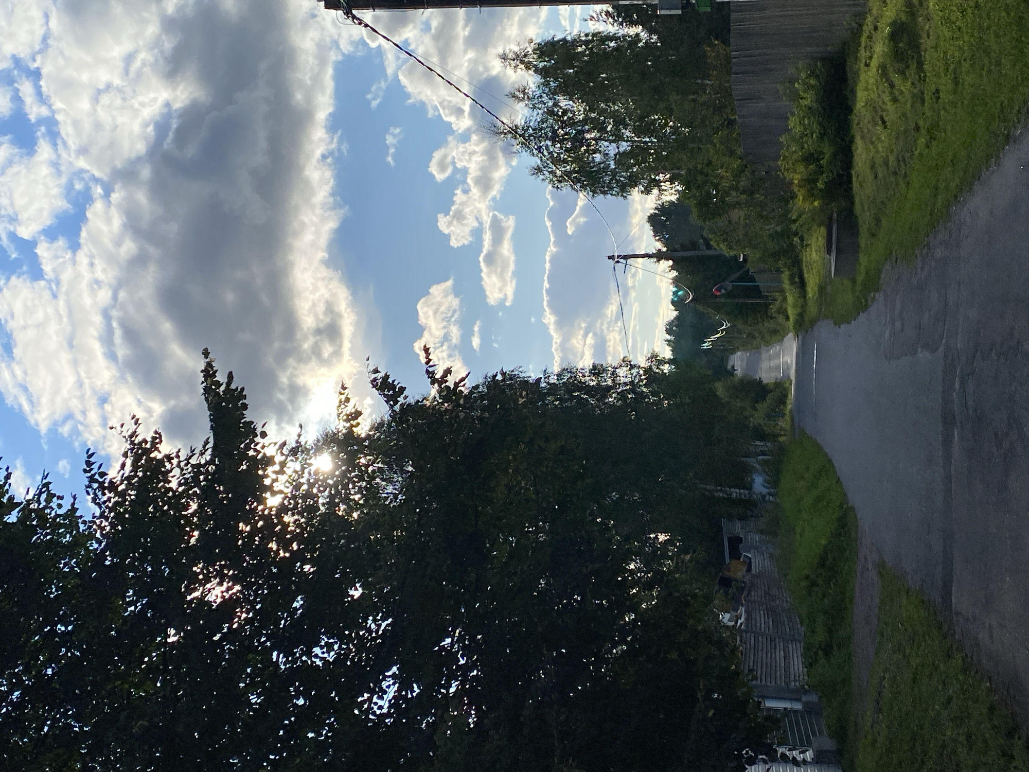 The image depicts a rural road in the village of Аист Снт, Russia, on a partly cloudy day. The road, paved with asphalt, winds uphill into the distance, bordered by grassy verges and wooden fences on either side. Dense foliage from trees, predominantly on the left, partially obscures the sun, creating dappled light on the road. In the background, more trees form a thick tree line, and power lines with poles are visible stretching across the scene. A traffic light, showing green and red, is positioned on the left side of the road further up. The overall impression is one of a quiet, rural setting with signs of human habitation and infrastructure.