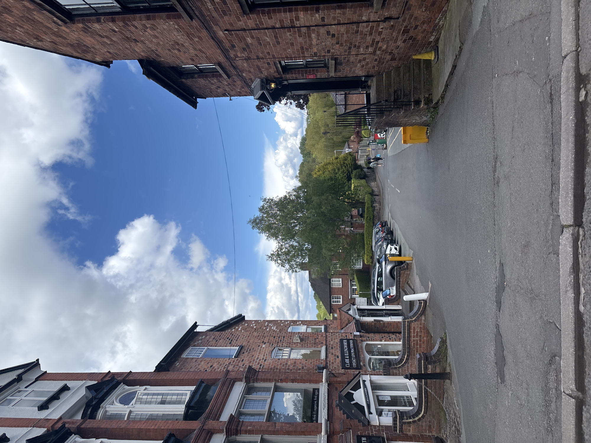 The image captures a street scene in Knutsford, United Kingdom. The street is flanked by brick buildings with storefronts, including one for Clare & Illingworth, a contact lens practitioner. Cars are parked along the side of the road. The sky is visible between the buildings, showing a mix of blue sky and white clouds. In the distance, greenery suggests a park or wooded area. People can be seen walking along the street. Street furniture includes yellow traffic cones and bollards. The road surface shows signs of wear and patching. The scene is captured at eye level and the photograph is shot at a vertical angle.