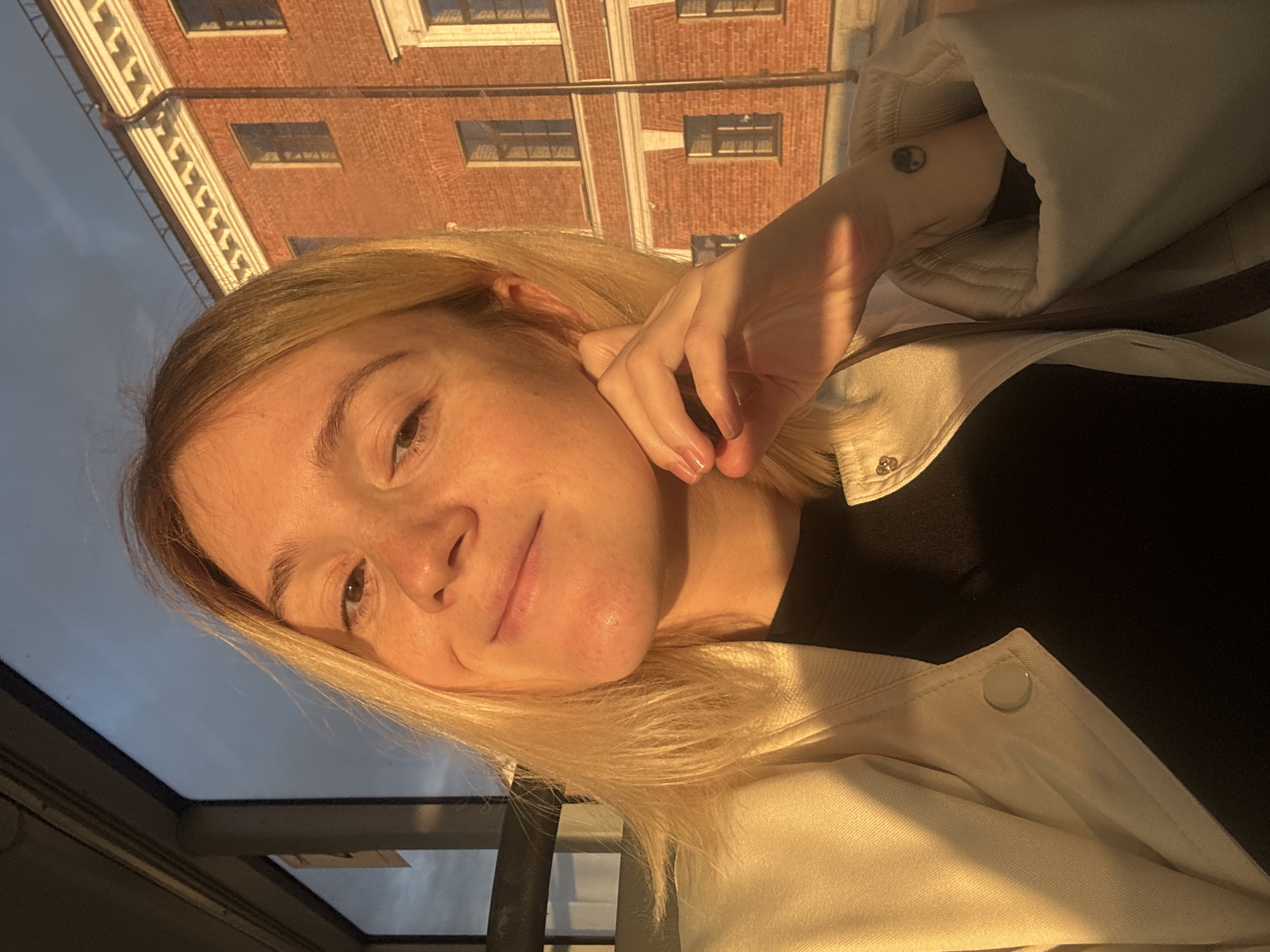 The image is a portrait of a young woman with blonde hair and fair skin, illuminated by the warm golden light of a sunset. She is looking towards the camera with a slight smile, her left hand resting near her face. She is wearing a light-colored jacket over a dark top. Behind her, a brick building with white trim and several windows is visible. The sky above is a mix of blue and grey clouds. A portion of a dark, industrial-looking structure with horizontal bars is visible on the left side of the frame, suggesting an outdoor setting. The specific location within Nizhny Novgorod, Russia, cannot be determined from the visual information present in the image.