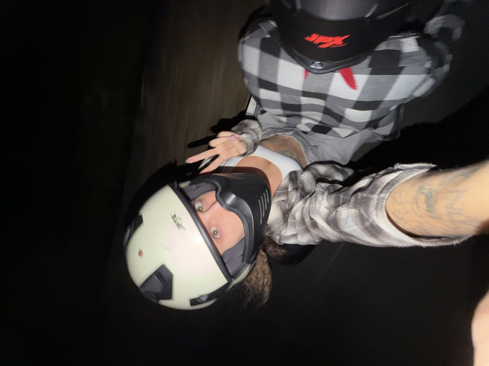 An angled, low-light selfie captures two individuals on what appears to be a motorcycle or scooter at night in North Kuta, Indonesia. The camera is held by the person in the foreground, whose face is partially visible through the open visor of a light-colored, cream and black full-face helmet. Their eyes are wide, looking directly at the camera, and their curly hair is visible beneath the helmet. This individual wears a black and white plaid long-sleeve shirt over a white crop top, and their left arm (right from the viewer's perspective) displays tattoos. With their right hand, they form a "peace" sign (V-sign).
Behind and slightly above them, a second person is visible, wearing a black full-face helmet with a red "JPX" logo on the forehead. This individual is also dressed in a black and white plaid shirt. The background is almost entirely dark and indistinct, suggesting the shot was taken at night or in very low light, possibly while in motion. The overall atmosphere is casual and likely playful, indicated by the selfie format and the hand gesture.