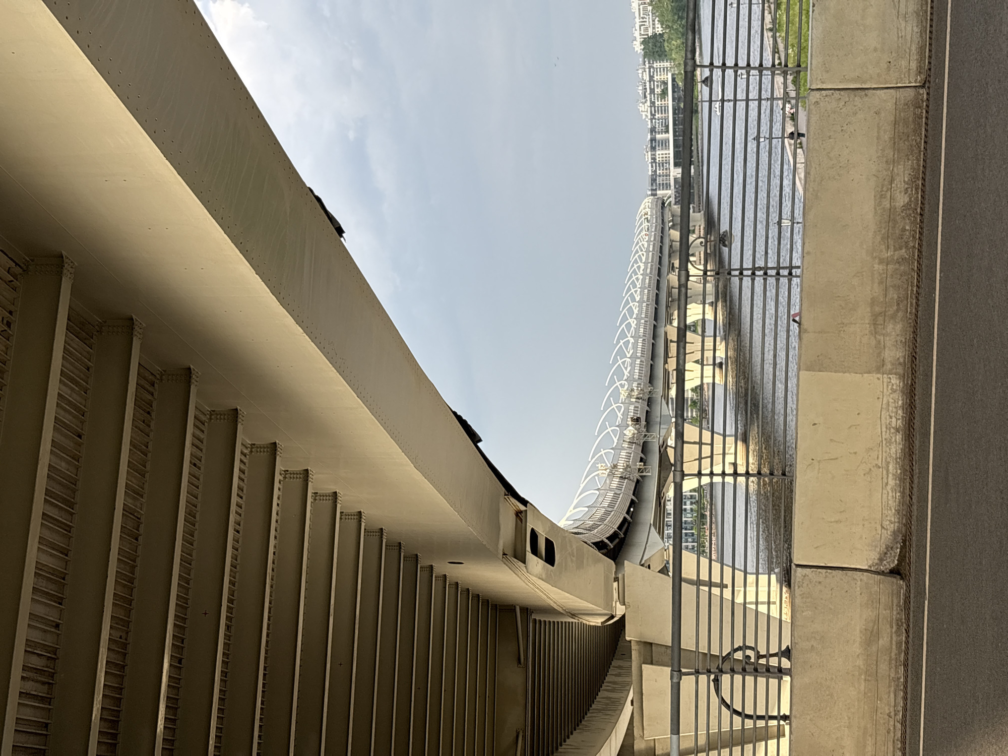 The image, originally rotated, captures a modern urban waterscape under bright daylight. A large, light-colored bridge or overpass structure dominates the left and central-left portions of the frame. This structure is characterized by a prominent curved profile, with numerous vertical support beams or ribs visible underneath, suggesting a robust metal construction. Its upper surface displays a line of rivets or fasteners and shows signs of weathering, including streaking. As it curves away, a rectangular opening or cutout is visible in its side.
Across the mid-ground, a body of water, likely a river or canal, stretches horizontally. A dark metal railing with vertical bars separates the water from a concrete-edged walkway or road on the immediate right. Several dark shapes, possibly birds or ducks, are visible on the water's surface and perched on the railing. On the opposite bank of the water, a dense array of multi-story urban buildings forms the background. Further along the waterway, another elegant bridge structure with a distinctive white, lattice-like design is visible, curving gracefully.
The sky above is bright and clear with some light clouds, indicating fair weather conditions during the day. No human activity or discernible text is visible. The architectural style points to a contemporary urban environment, consistent with modern infrastructure found in Saint Petersburg, Russia.