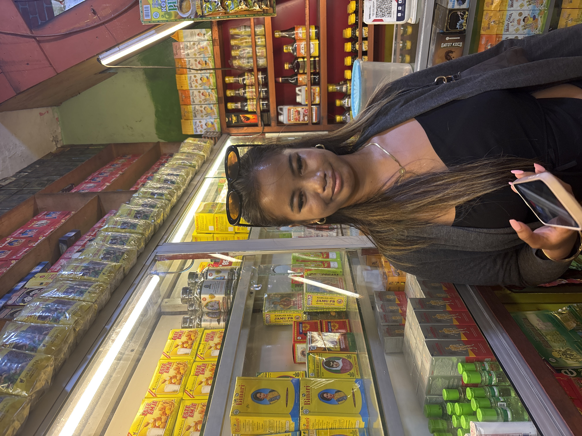 The image depicts a person standing in front of a display of various products, possibly in a store. There are rows of items including boxes of Binari and Jamu PB products, as well as Tolak Angin and Este Emje products. There are also bottled liquids on the shelves. The person is wearing a gray jacket over a black top and has sunglasses perched on their head. The location may be in North Jakarta, Indonesia, considering the local language on the products. The person holds a smartphone.