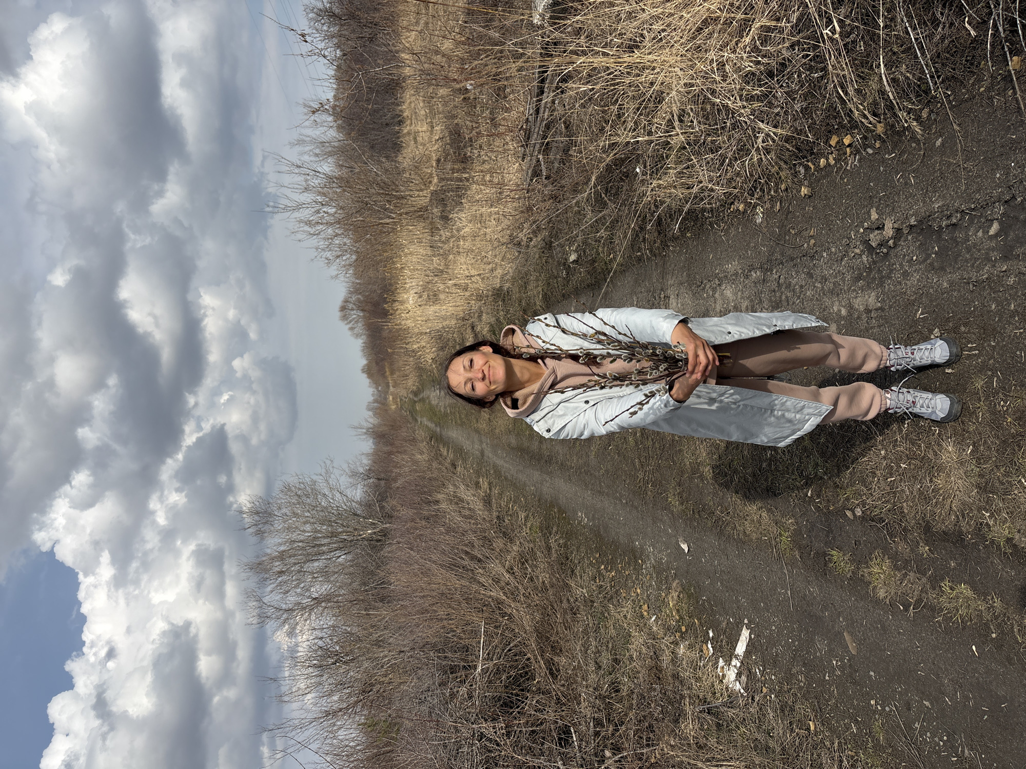 A woman is standing on a dirt path in Ulyanovsk, Russia. She is holding branches with furry buds. The path is flanked by dry vegetation. A cloudy sky is visible in the background. The woman is wearing a light-colored jacket, beige pants, and gray sneakers.