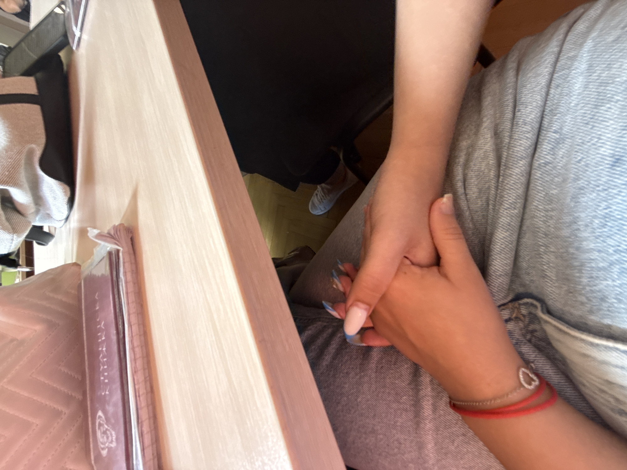 The image shows a close-up view of two pairs of hands clasped together under a light-colored wooden desk. One person's hands are resting on their lap, which is covered in blue denim jeans. A red string bracelet with a small silver heart charm is visible on the wrist of this person. The other person's hands are intertwined with the first, and they have long, manicured nails painted with a French tip and blue accents. Above the desk, a light pink quilted object and a dark red booklet with Cyrillic text are visible. In the background, a light gray bag with black straps is on the desk, and a person's legs, wearing light gray sneakers and black pants, are visible under the desk. The scene appears to be an indoor setting, possibly a classroom or office, in Sofia, Bulgaria, given the context provided.