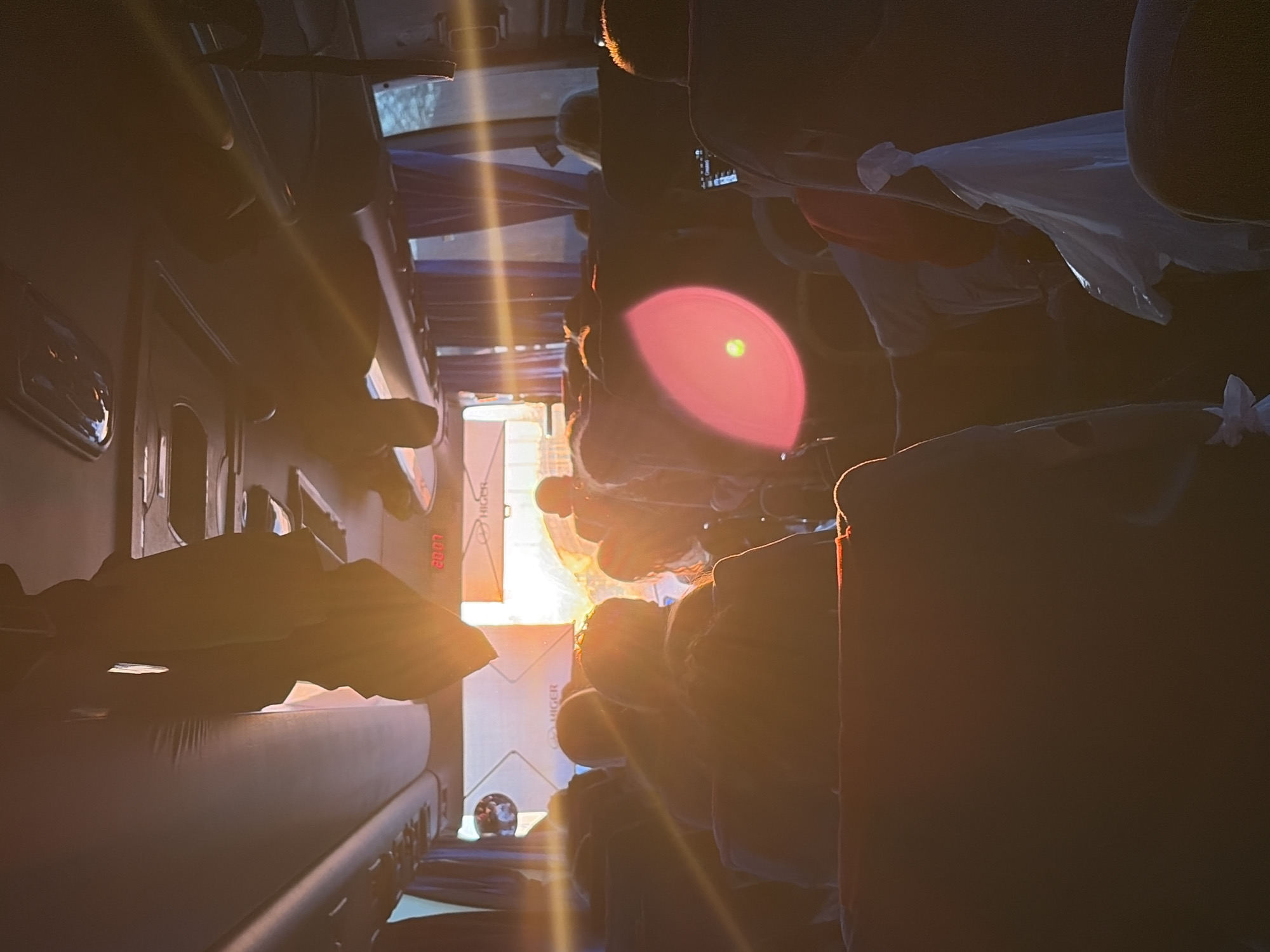 The image shows the interior of a bus with multiple rows of seats, most of which appear to be occupied. Sunlight is streaming through the front of the bus, creating a bright glare that obscures some details. A luggage rack is visible on the left side, and the word "HIGER" appears on a panel near the front. A small globe-like object rests on the floor beneath the luggage rack. The image is strongly lit and slightly disorienting, likely taken from a low angle. There is no visual evidence confirming the location to be Ustinovo, Russia.