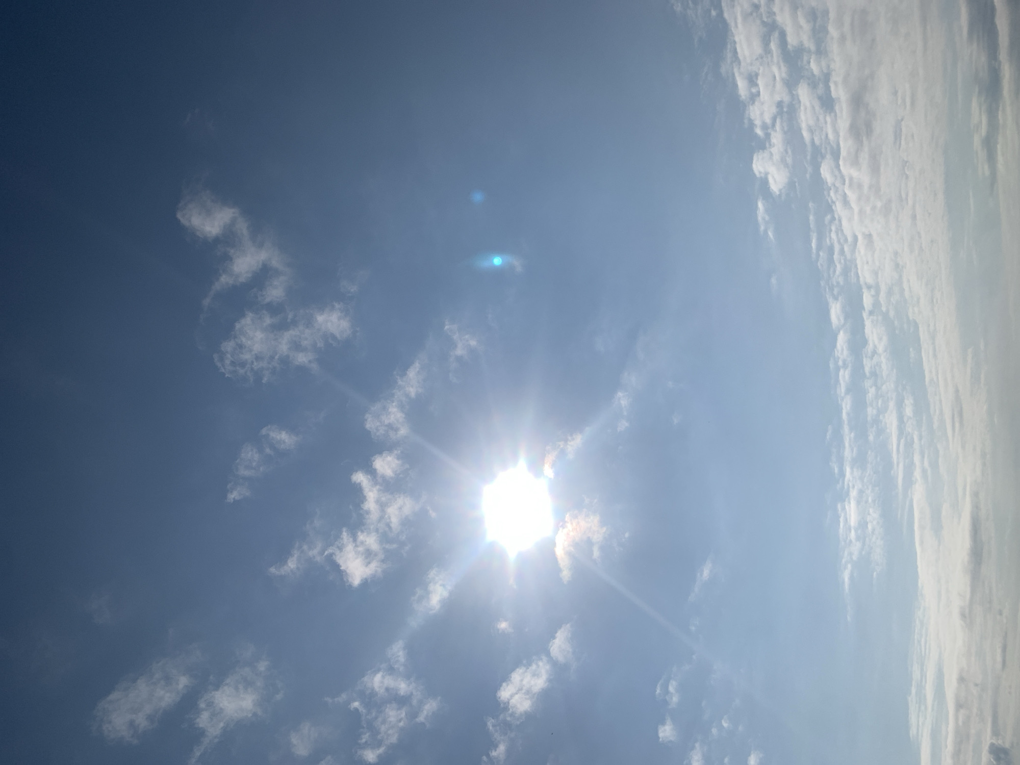 The image shows a bright sun shining in a clear blue sky with scattered white clouds. The sun is a prominent feature, with rays of light emanating from it. There are some lens flares visible in the sky, one appearing as a bluish-green orb and another smaller one to its right. The clouds are wispy and appear to be at various altitudes. The bottom of the image shows a layer of denser, whiter clouds, possibly indicating a horizon or lower atmospheric layers. The overall scene depicts a sunny day. No specific location in Kolomyia, Ukraine can be determined from the image. There are no people, objects, or actions visible.