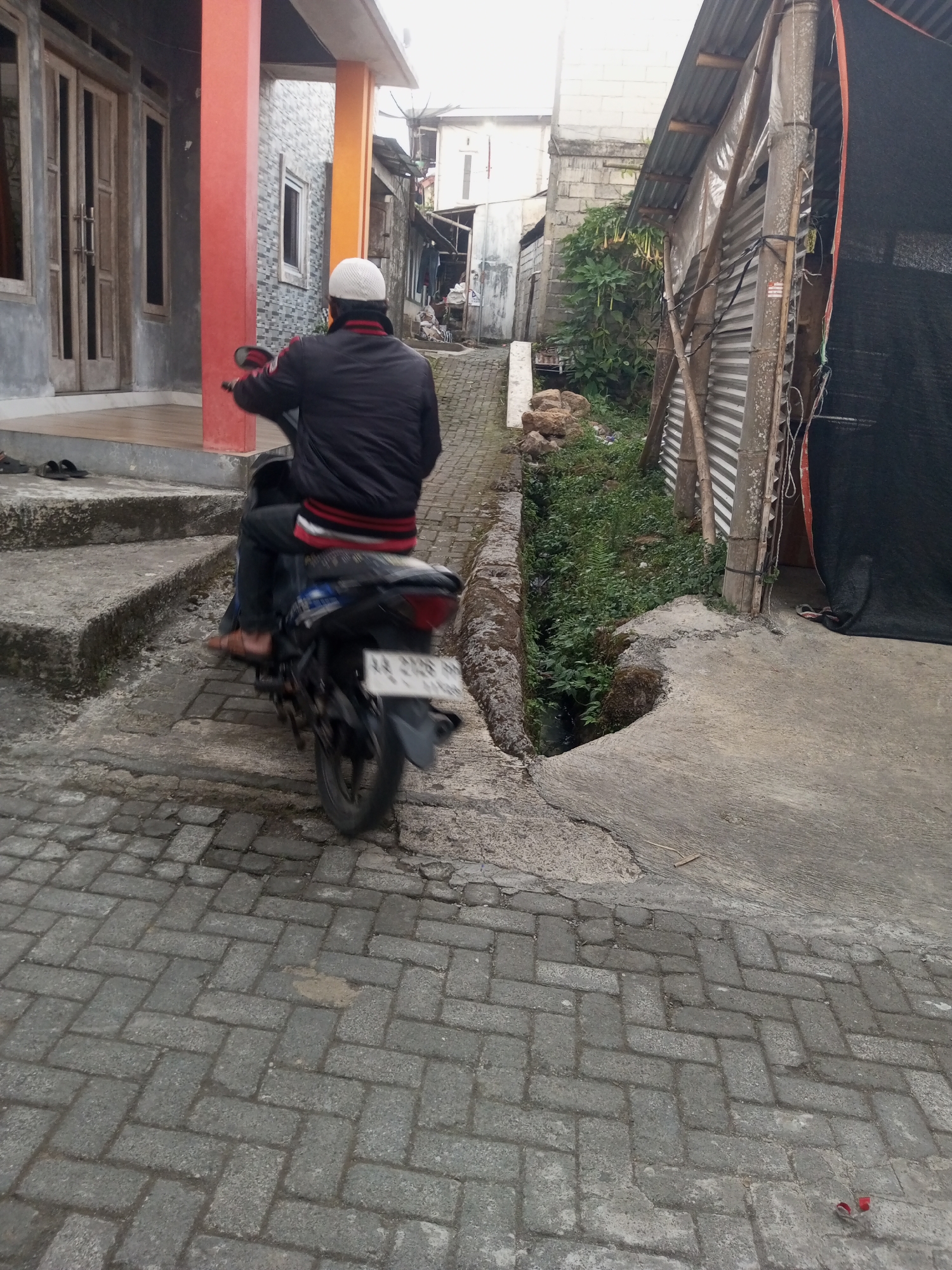 The image shows a view of a narrow street in Andongsili, Indonesia. A person is riding a motorcycle away from the camera. The person is wearing a black jacket with red and white stripes on the sleeves and a white cap. The motorcycle has a license plate that is mostly obscured by dirt and motion blur. The street is paved with cobblestones and bordered by a concrete drainage ditch on the right. On the left side of the street, there is a house with red and orange pillars and a wooden double door. The background shows more houses and vegetation. The overall impression is of a humble urban or semi-urban environment.