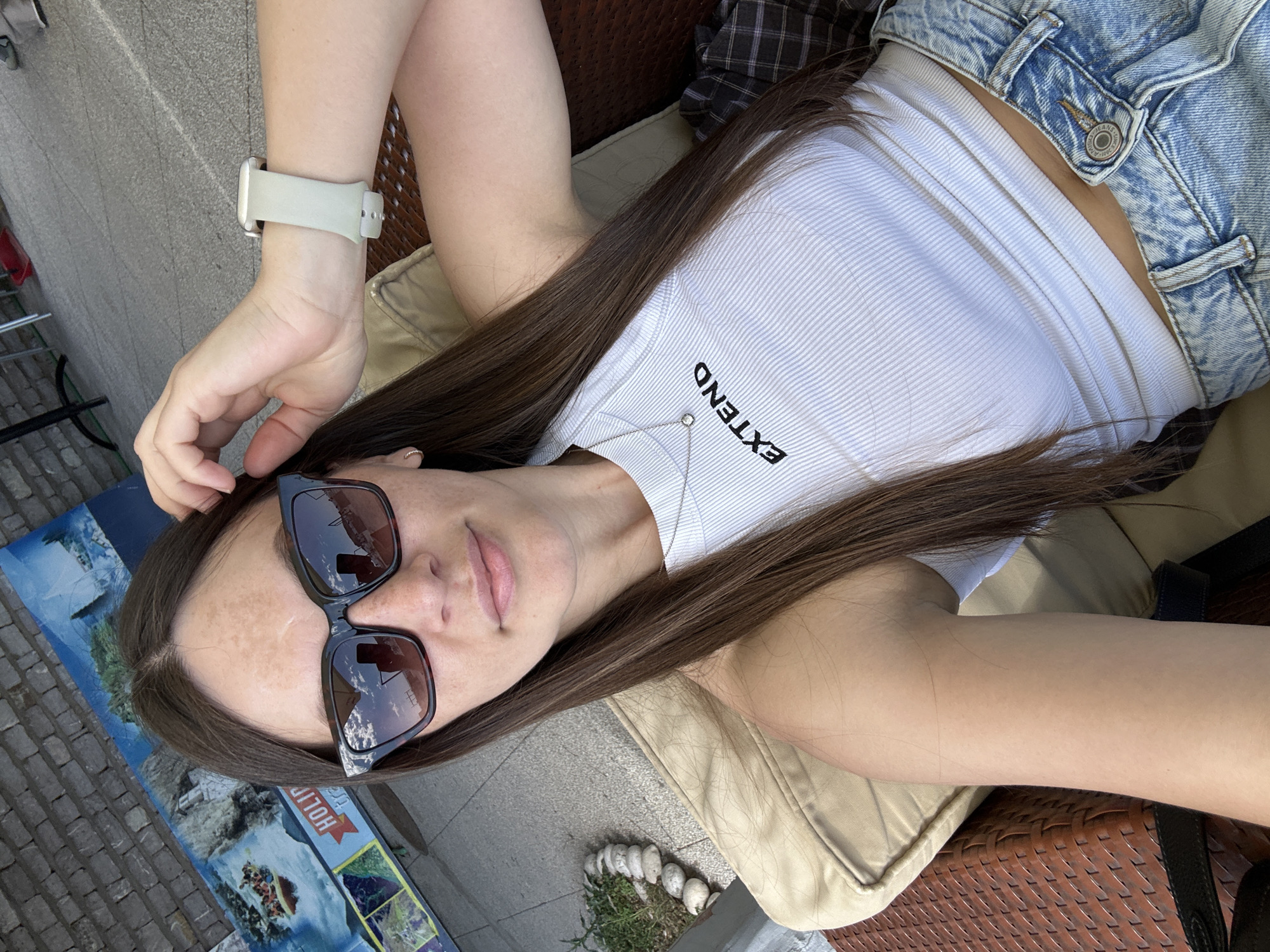 A selfie of a young woman is the central focus, partially reclined and appearing to be seated. She has long, dark hair, fair skin, and wears dark sunglasses. Her attire consists of a white ribbed top and light-wash denim shorts. On her left wrist, she wears a light-colored smartwatch. The woman's body is at an angle, with her head and torso visible in the frame. The word "EXTEND" is printed vertically on her shirt.
She rests on what appears to be a tan cushion, with a woven chair visible beneath. A folded plaid garment sits beside her. Below, on the left side, is a promotional advertisement featuring images of water rafting. Cobblestone paving is visible in the background.
The location is likely Budva, Montenegro, inferred from the tourism advertisement depicting rafting activities that are common in the region.