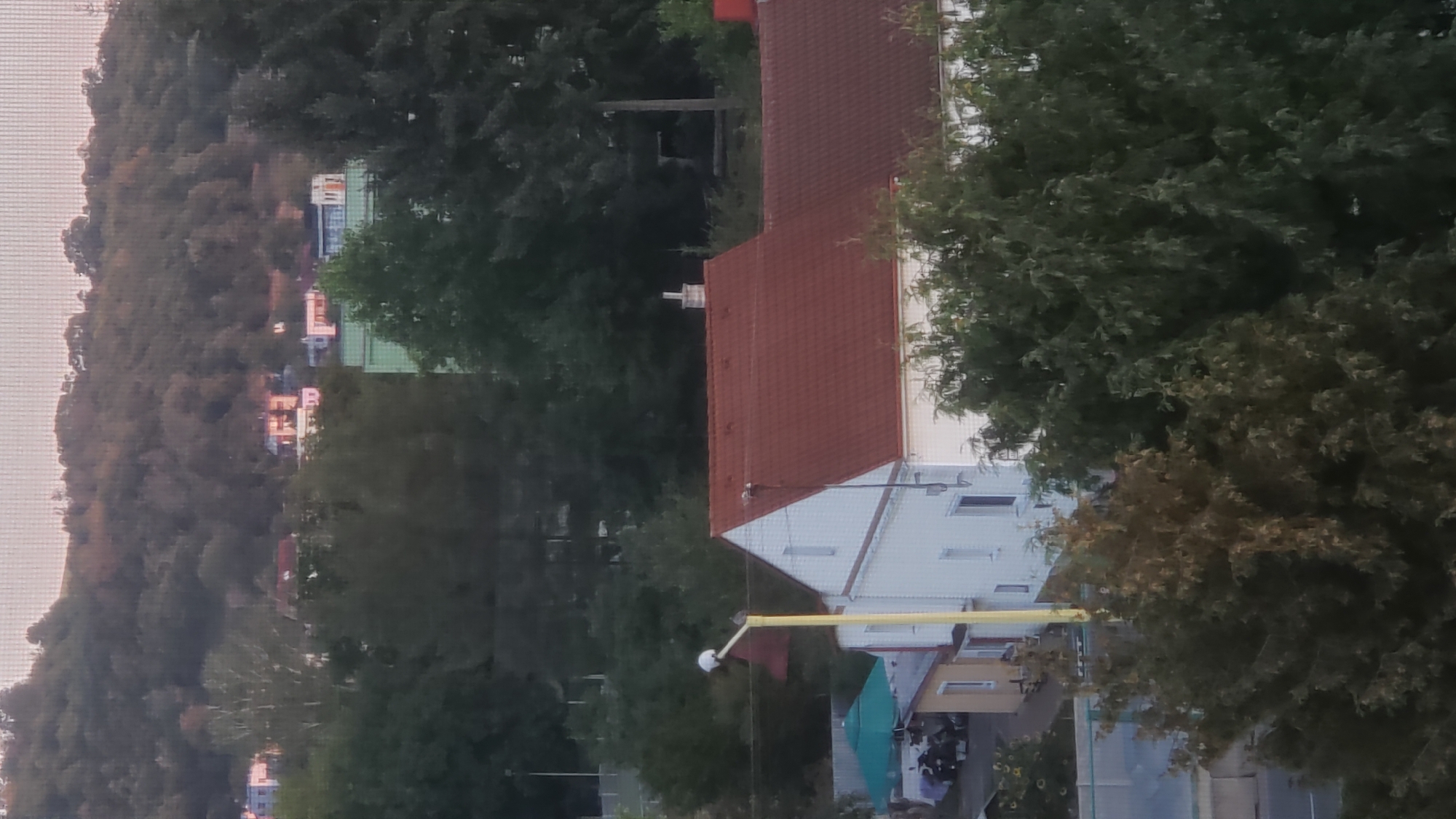 The image shows a residential area in Prešov, Slovakia, as seen from an elevated perspective. In the foreground, a large house with a red tile roof is visible, with a white exterior and several windows. To the left of this house, a smaller yellow building with a white trim is situated, and next to it, a green canopy is extended over an area with parked vehicles. A tall yellow pole with a light fixture and a red flag is positioned in front of the yellow building. Behind these structures, a dense line of trees forms a mid-ground. In the background, a hilly landscape is covered with vegetation, and several buildings with varying colors and roof types are scattered across the slopes. The sky above is a pale, hazy color, suggesting it might be dawn or dusk. The overall impression is of a quiet suburban or semi-urban environment.