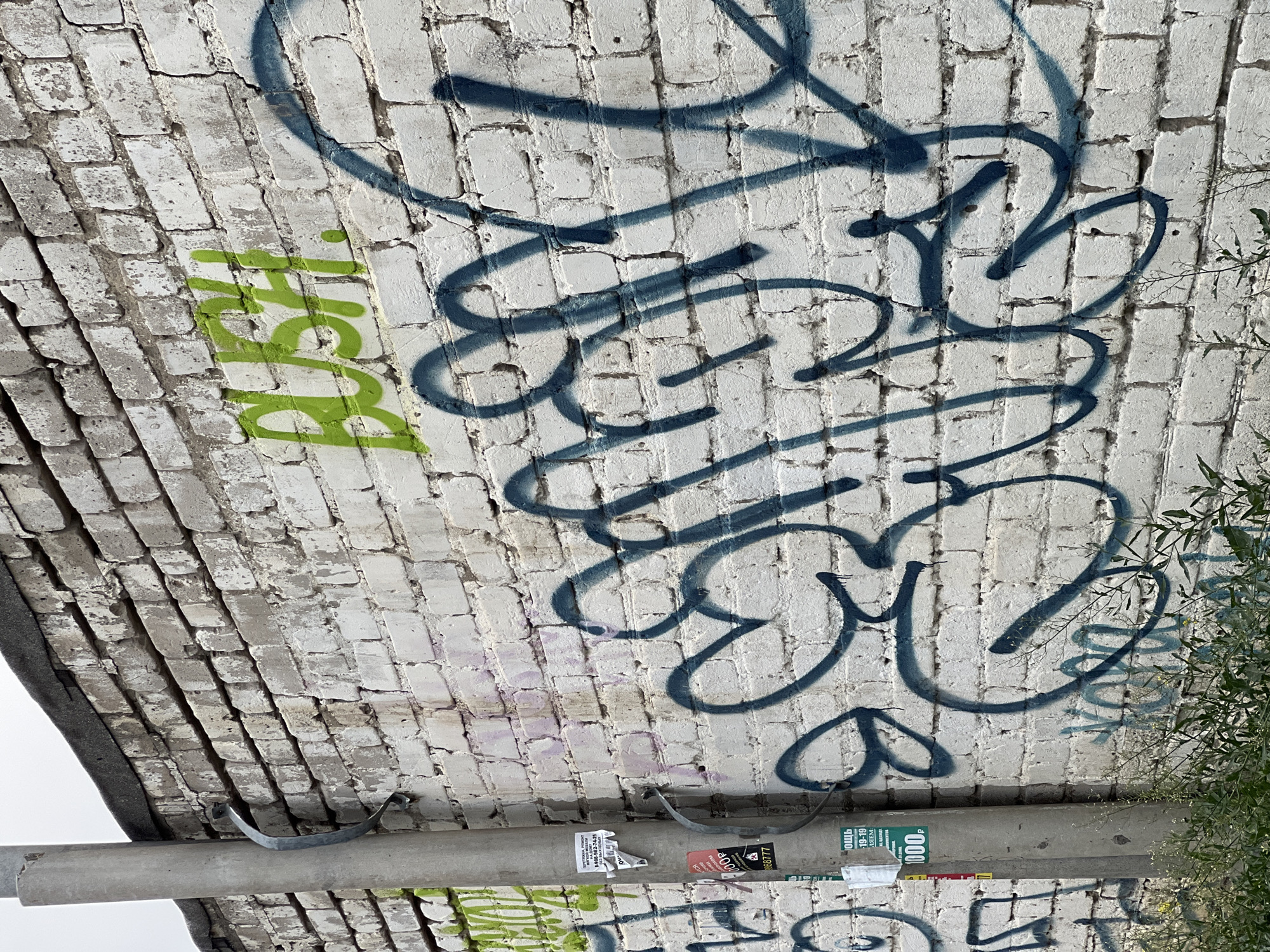 A low-angle, close-up photograph shows graffiti on a white brick structure in the city of Unknown, Unknown. The structure appears to be an arched brick ceiling or underpass, with a gray, cylindrical support beam running horizontally at the bottom. The graffiti consists of a larger, stylized tag in dark blue, and a smaller, vertical tag in green. The support beam is partially obscured by various stickers and remnants of adhesives. Vegetation is visible along the right edge of the image, partially obscuring the graffiti. The lighting is diffuse, casting soft shadows on the brick surface.