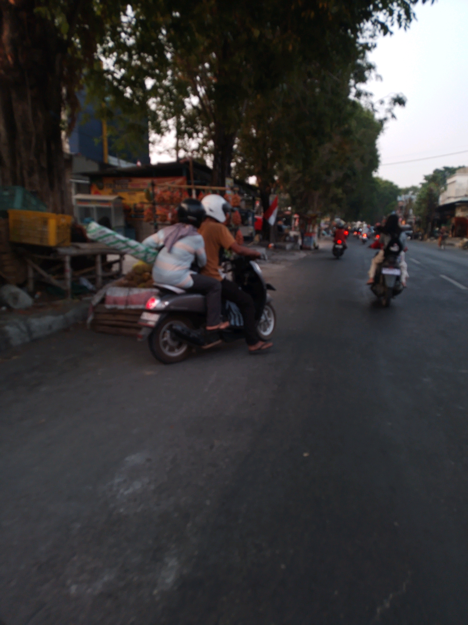 The image depicts a street scene in Kertosono, Indonesia, characterized by a low-angle view focusing on a motorcycle carrying two people. The motorcycle is a dark color, possibly gray or black, with visible wheels and a rear light. The front rider is wearing a dark helmet and a long-sleeved shirt, while the passenger behind them is wearing a light-colored long-sleeved shirt and a head covering.
To the left of the motorcycle, a wooden table is laden with what appears to be produce, possibly durian fruit, as well as some packaging. A yellow crate sits on the ground nearby. Behind these objects, a building with a blue facade and a dark roof is partially visible, suggesting a commercial or residential area.
The street is paved and appears to be a typical urban thoroughfare, with trees lining the background. Other motorcycles are present further down the street, some with riders, indicating ongoing traffic. The overall lighting suggests it is late afternoon or early evening, with the sun casting shadows and the sky appearing overcast. The image is somewhat blurry, suggesting motion during the capture or a low-resolution camera.