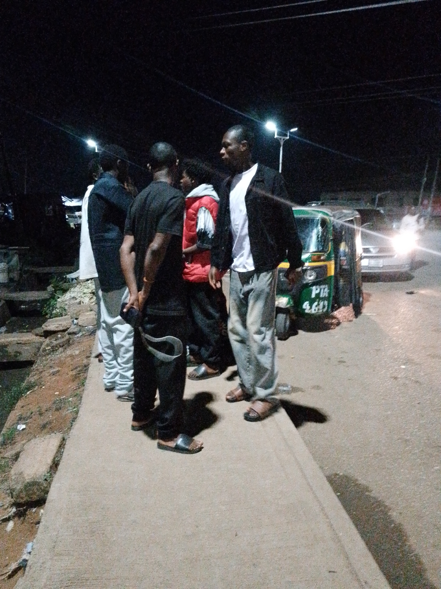 The image depicts a nighttime scene in what appears to be an urban or semi-urban environment. Several individuals are standing on a paved walkway next to a road. In the background, a green and yellow auto-rickshaw is parked, and a car with its headlights on is visible. The overall lighting suggests it is dark, with streetlights providing some illumination.
Focusing on the people, there are at least five individuals visible.
One man in the foreground, facing away from the camera, is wearing a black t-shirt and dark pants. He has a light-colored strap around his waist. His right hand is holding a dark object. He is wearing dark sandals.
To his left, another man is wearing a black jacket over a white t-shirt and light-colored pants. He is facing towards the auto-rickshaw and appears to be looking in that direction. He is wearing brown sandals.
Further to the left, partially obscured, are two more individuals. One is wearing a white shirt and light pants. The other is wearing a red vest or jacket over a darker garment.
Another individual is visible further to the left, wearing a dark jacket over a white shirt and light pants.
The environment includes a concrete walkway, a dirt or gravel area to the left with some vegetation, and a paved road to the right. The auto-rickshaw has markings on its side, including what appears to be "PTA 4610". The car in the background has its high beams illuminated. The scene conveys a sense of a roadside gathering at night.