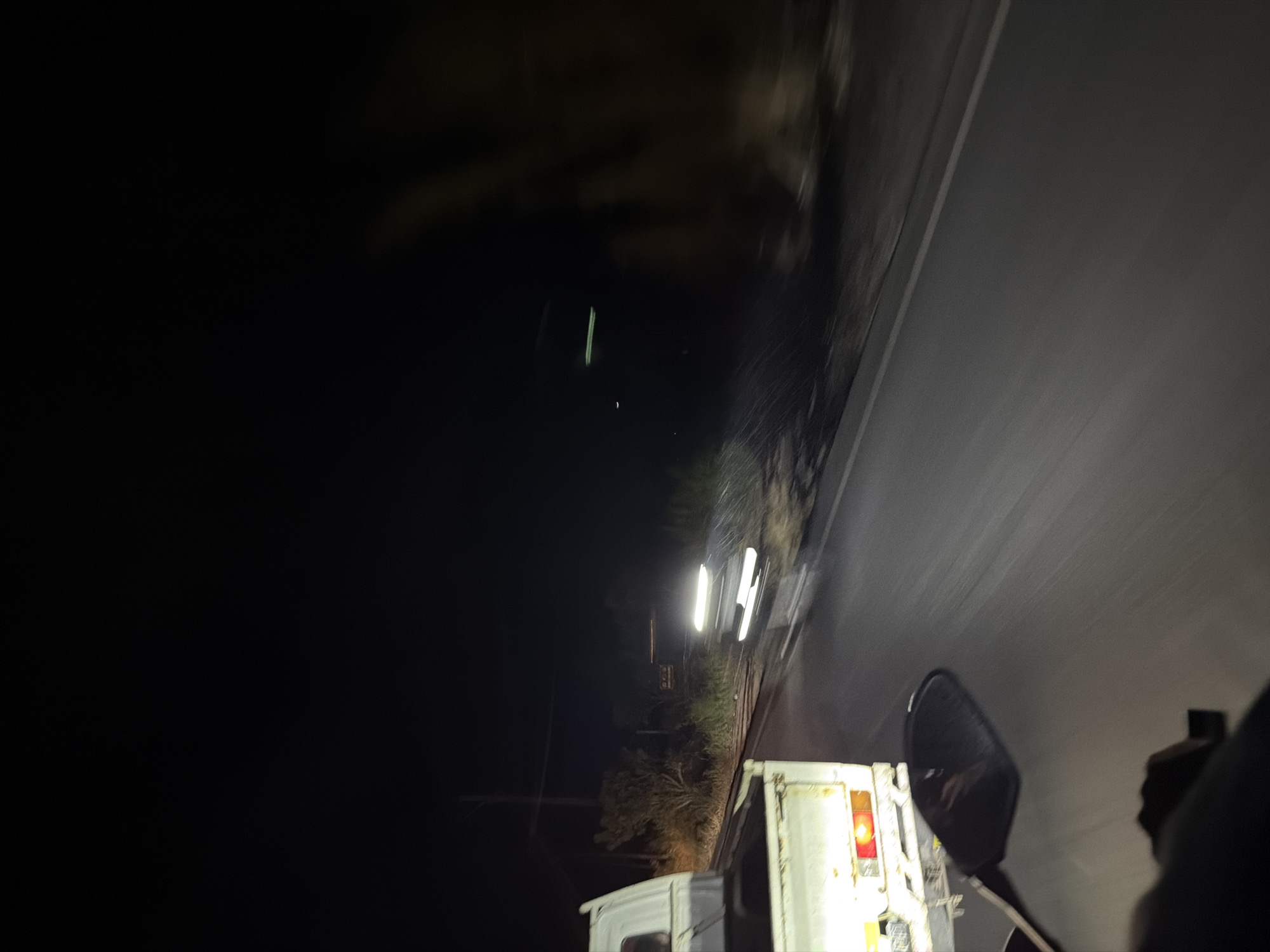 The image captures a nighttime scene on a road, likely in Kintamani, Indonesia, characterized by dark surroundings and illuminated by vehicle headlights. In the foreground, the rear of a white pickup truck is visible on the left, with its brake lights illuminated. A motorcycle's side mirror is also in the lower left quadrant of the frame. Further down the road, another vehicle's headlights are clearly visible, creating a bright, blurred streak due to motion. The environment appears to be a rural or semi-rural area, with vegetation and a dark sky suggesting the absence of significant urban lighting. The road surface is asphalt and shows signs of being wet, reflecting the lights. The overall impression is one of travel at night, with the blurred elements indicating movement.