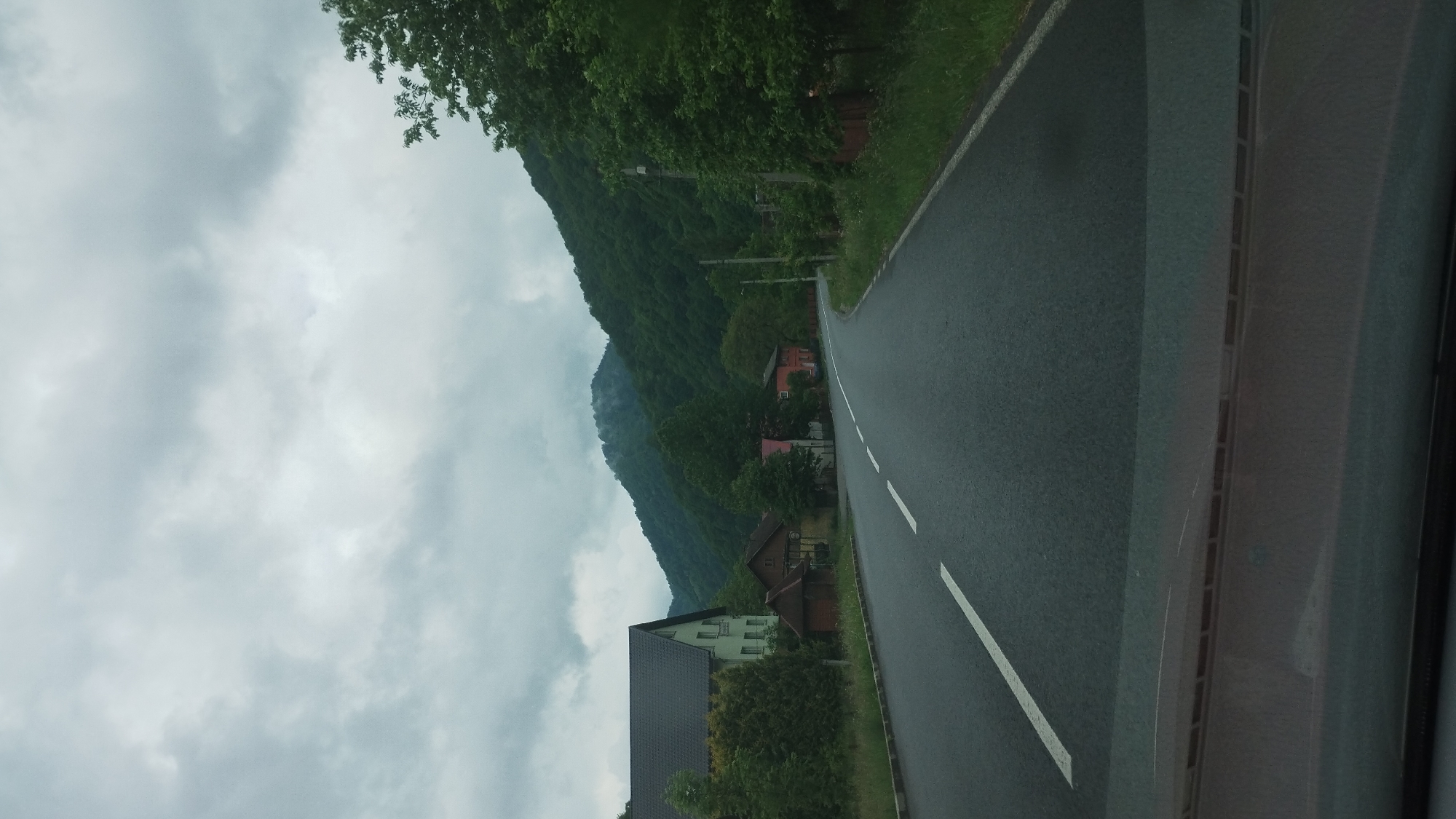 The image shows a view from a vehicle driving on a wet, paved road in Bilyn, Ukraine. The road has solid white lines indicating the lane markers. To the left of the road, a steep, tree-covered mountain slopes upwards, disappearing into a cloudy sky. Several houses with red and green roofs are nestled among the trees on the mountainside. The sky is overcast with thick, grey clouds, suggesting it is either a cloudy day or has recently rained. The overall atmosphere is somewhat subdued due to the weather. The image appears to have been taken in the daytime, but the cloudy conditions make it difficult to determine the exact time. There is no visible text in the image.