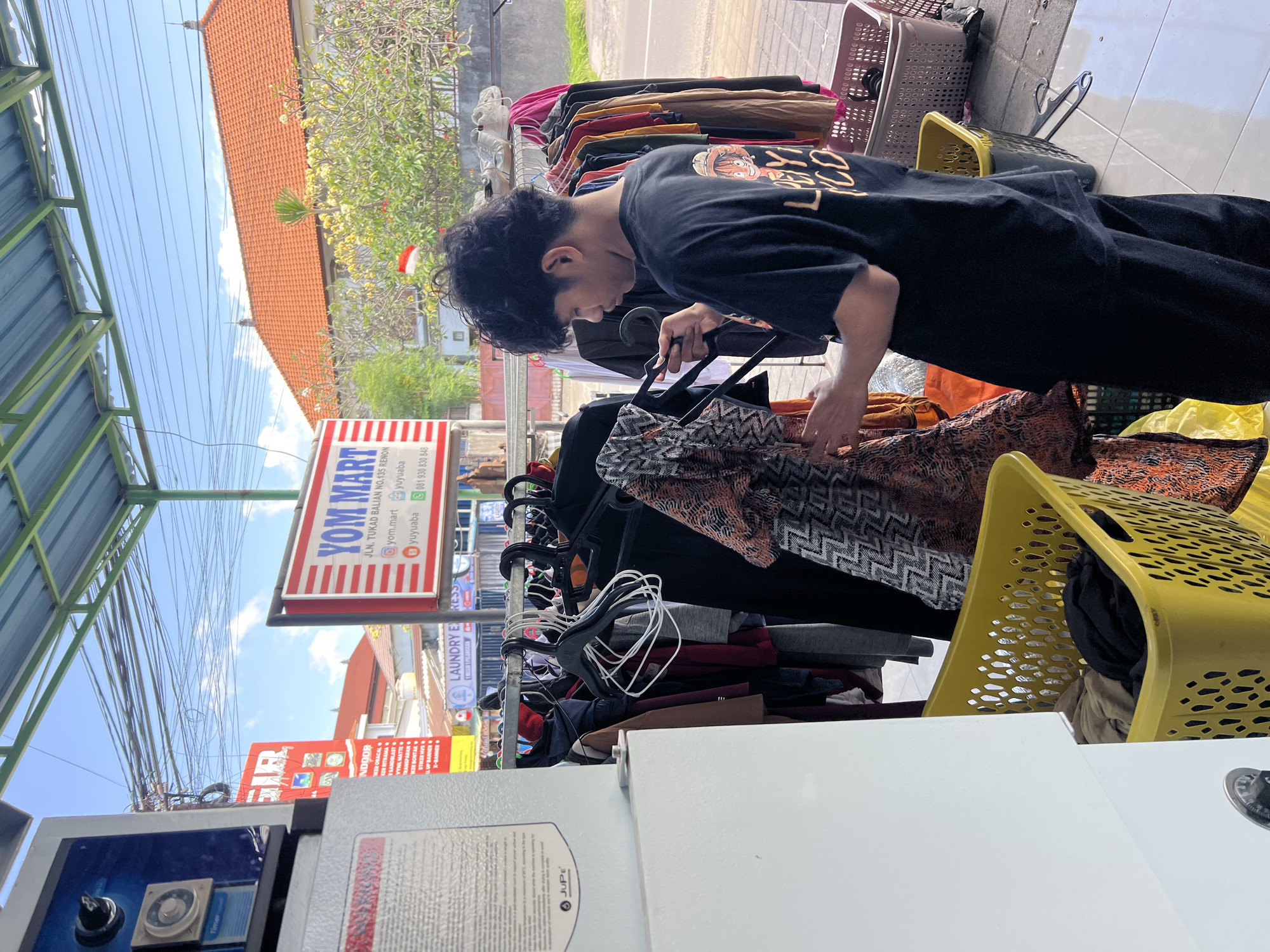 A young man with curly dark hair, wearing a black t-shirt with a cartoon image on the back and black pants, is standing on a tiled sidewalk outside a laundry service. He is holding a garment with an orange and black pattern and appears to be inspecting it or preparing it for washing. Behind him, clothes hang on a rack, and several laundry baskets, one yellow and one pink, are visible. A sign for "YOM MART" is mounted on the wall, and a red and white sign advertising printing services is also present. The environment suggests a commercial area in Denpasar, Indonesia, with buildings and a street visible in the background. The sky is blue with some clouds.