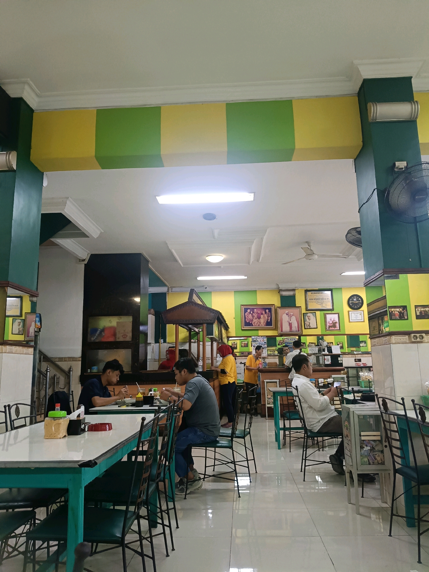 The image depicts the interior of a restaurant, likely located in Dapur, Indonesia, characterized by its vibrant green and yellow decor. In the foreground, two men are seated at a white-topped table, engaged in eating. Their table is equipped with condiment dispensers and a red tray. The seating arrangement includes dark green chairs with black metal frames. Further into the restaurant, several individuals are visible, including two women in yellow tops and another person in a red headscarf, who appear to be working behind a counter. A ceiling fan is visible, along with fluorescent lighting. The walls are adorned with framed photographs and what appears to be a clock. The floor is tiled with a glossy finish. The overall ambiance suggests a casual dining establishment.