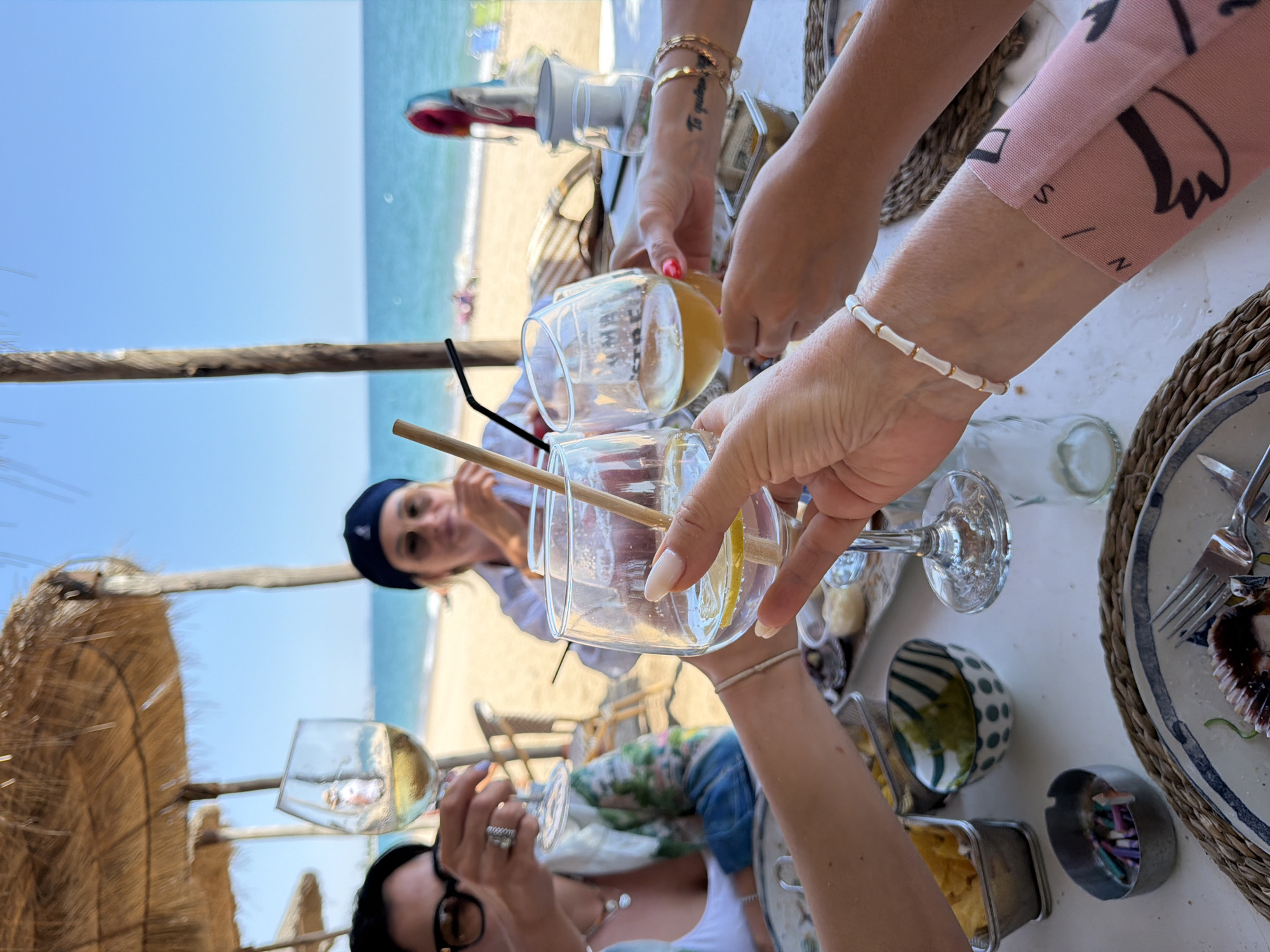 The image captures a sunny, beachside setting in Mont-roig del Camp, Spain. A group of people is enjoying a meal at a table under a thatched-roof structure. The beach and ocean are visible in the background. A woman in a blue hat is in the center of the image. Two people are pouring drinks. Plates, silverware, bowls, and drinkware are on the table. The focus is on a shared meal and drinks among friends.