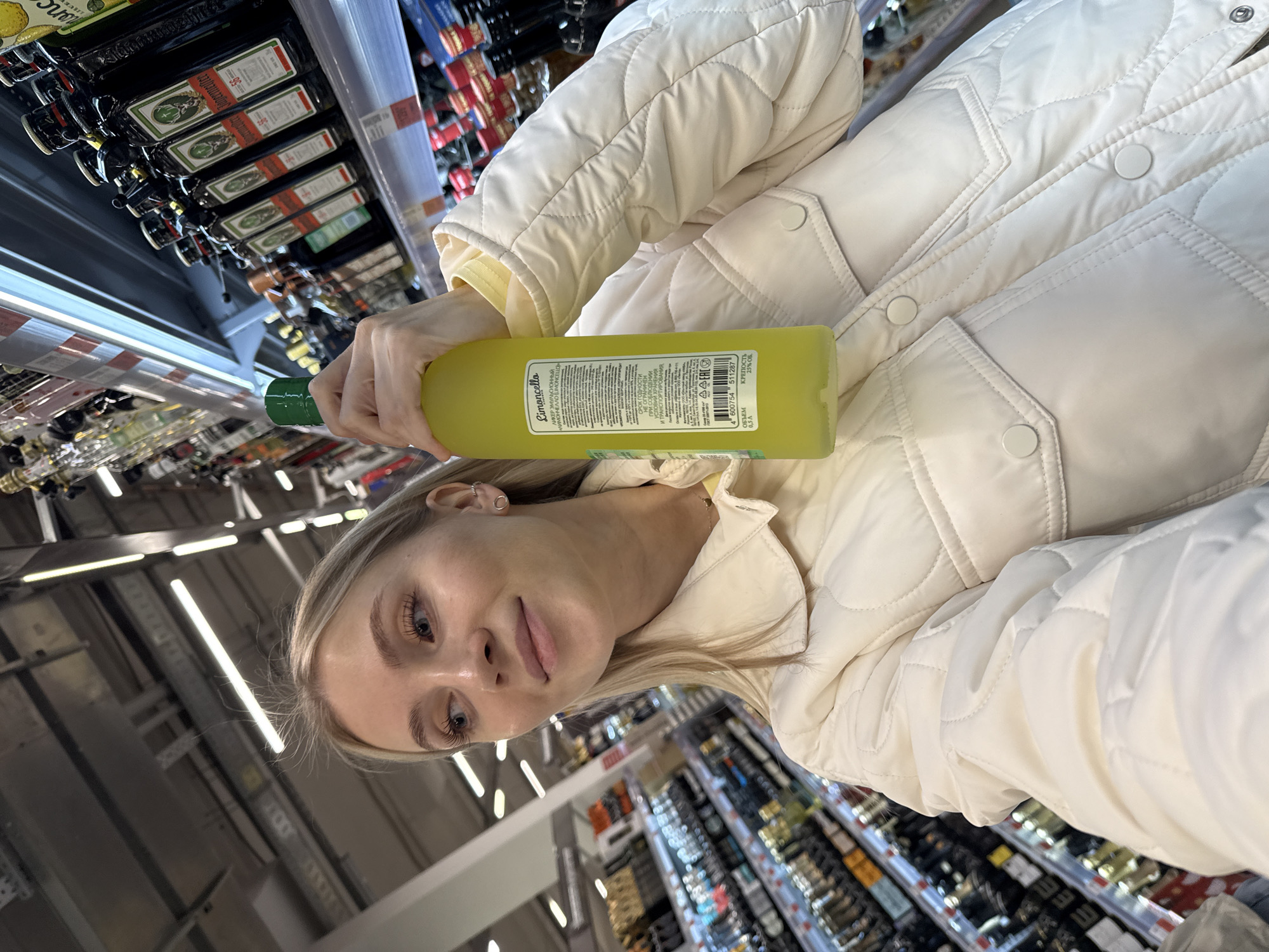 A woman is holding a bottle of Limoncello liqueur while standing in the alcohol aisle of a supermarket. The bottle is green and yellow, with Russian text on the label. The woman has blonde hair and is wearing a white jacket. The supermarket aisle is stocked with various alcoholic beverages, including Jägermeister. The photo was likely taken in a supermarket in Izhevsk, Russia, based on the Cyrillic text on the Limoncello bottle.