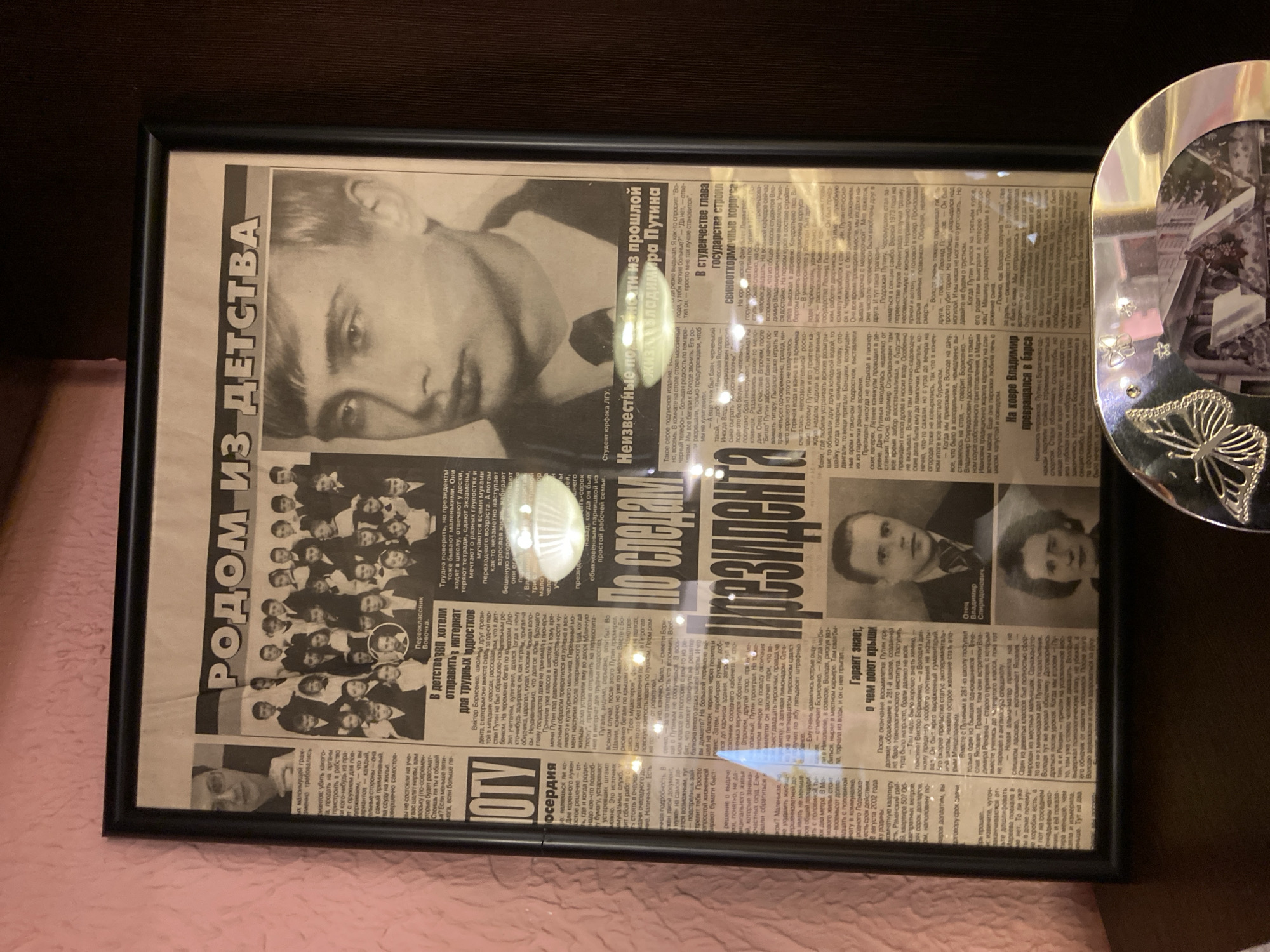 The image shows a framed newspaper clipping. The clipping features black and white images, including what appears to be a young Vladimir Putin. The headline translates roughly to "From Childhood." Other text and images on the clipping are in Russian. The frame is black. The newspaper clipping is positioned against a dark brown background, possibly a wall or furniture. To the right of the frame is a decorative heart-shaped object with a butterfly emblem. This image was likely taken in Sergiyev Posad, Russia.