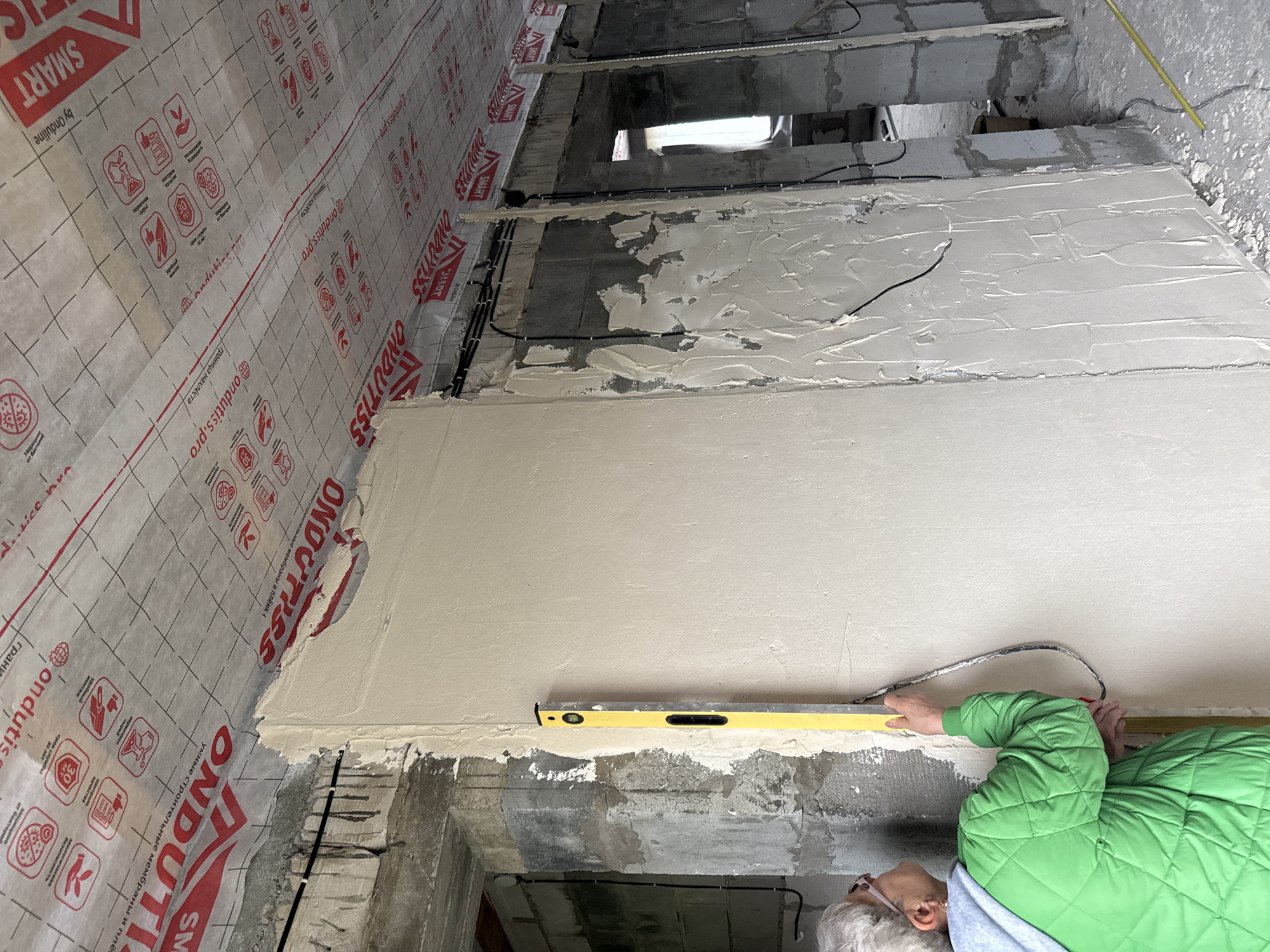 In an indoor construction setting, a person wearing a bright green puffer jacket and glasses is using a yellow spirit level to check the flatness of a newly plastered wall. The wall has a smooth, light-colored finish, and the spirit level is positioned horizontally against it. The background shows unfinished concrete structures, exposed wiring, and building materials. The walls are partially covered with a white and red patterned membrane, likely a construction vapor barrier, with visible text such as "SMART," "Ondutis," and "ondutiss.pro." The overall scene suggests a renovation or construction project in progress. The lighting indicates it is daytime, but the specific time of day and weather conditions are not discernible. The location is identified as Perm, Russia.