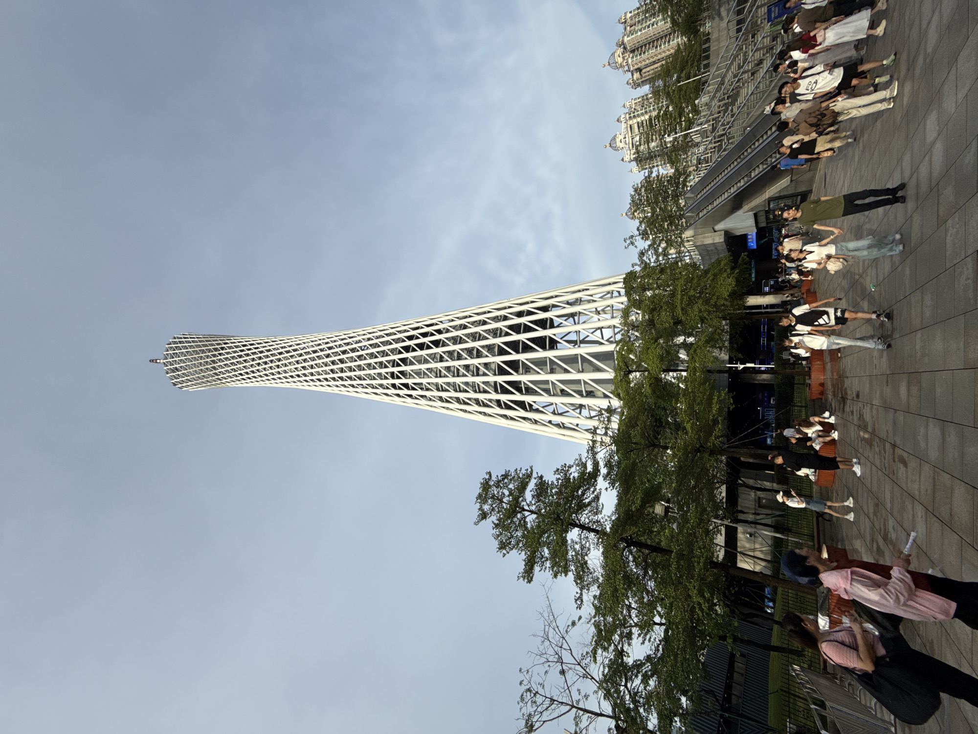 In Haizhu District, China, a group of people are gathered in a public space near the Canton Tower. The tower, a tall, latticed structure, dominates the background. Trees line the paved area where pedestrians are walking and standing. Several buildings with distinctive domed roofs are visible in the distance. An escalator leads up to a higher level of the plaza. The scene appears to be set during the daytime, with a clear sky.