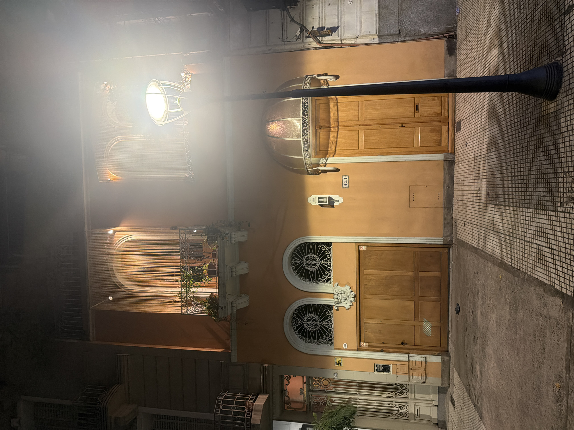 The image depicts a street scene at night in Buenos Aires, Argentina. A two-story peach-colored building is illuminated by a streetlamp. The building has two main entrances, each with a wooden door. The upper entrance features an arched awning with decorative ironwork. The lower entrance is flanked by circular windows with ornamental grilles. Windows on the upper floor are adorned with string curtains and plants. The sidewalk is paved with mosaic tiles. A black streetlamp is positioned in the foreground.