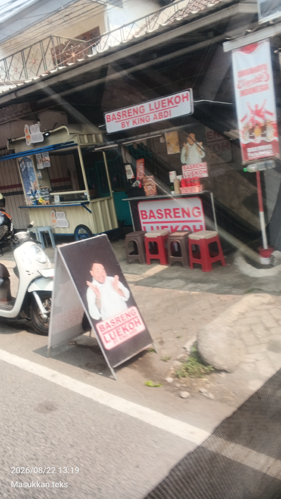 In the city of Pakis, Indonesia, a street-side food stall displays a sign reading "BASRENG LUEKOH BY KING ABDI." A white scooter is parked to the left of the stall, and a man is pictured on an A-frame sign in front of the stall. The stall is situated along a road, with several small red and brown stools placed in front of it. A white banner with red lettering hangs from the building behind the stall, and another red and white banner is mounted on a pole to the right. The scene is illuminated by sunlight, casting shadows on the ground. The date and time stamp "2026/08/22 13:19" and the text "Masukkan teks" are superimposed on the lower left of the image.