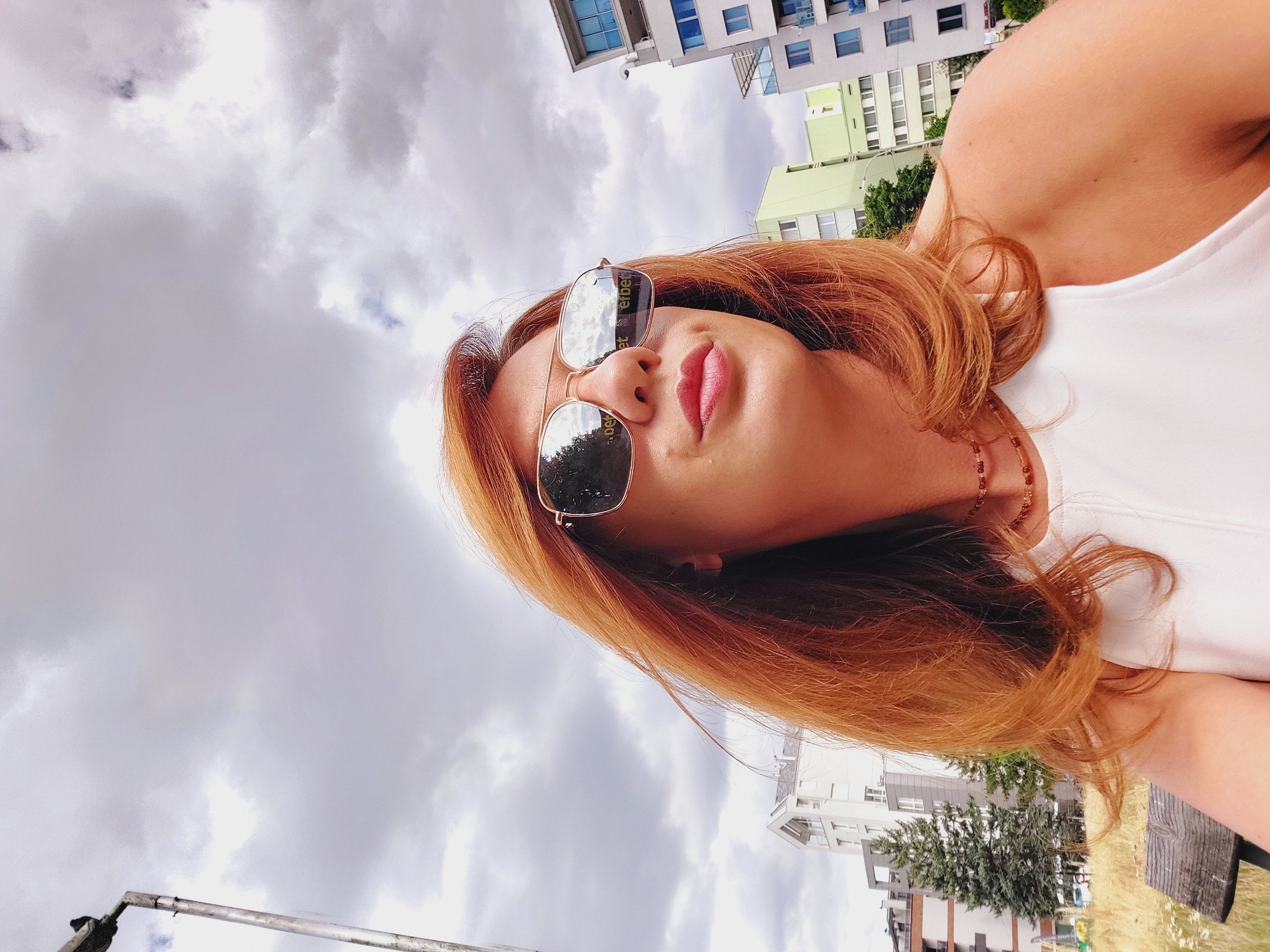 A woman with auburn hair, wearing sunglasses and a white top, is captured in a low-angle selfie outdoors. The sunglasses reflect buildings and possibly a cloudy sky. In the background, modern apartment buildings of various colors, including white, light green, and blue, are visible. A lamppost stands to the left, and a wooden bench is partially visible in the lower left corner. The setting appears to be an urban environment, likely in Sofia, Bulgaria, given the architectural style and the presence of trees and greenery. The sky is overcast with visible clouds.