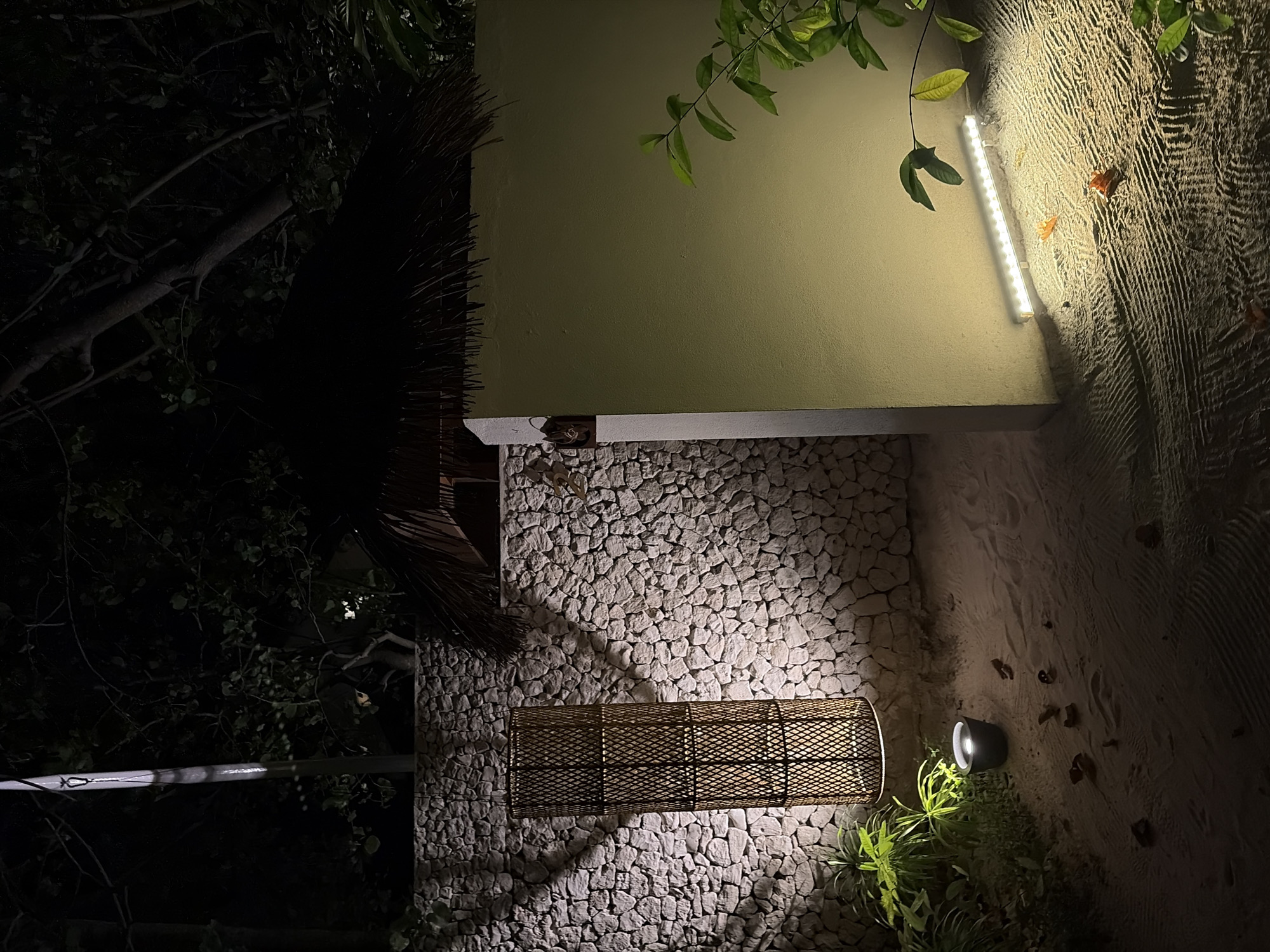 In the city of Halaveli, Maldives, an outdoor scene is illuminated by artificial lighting. The foreground consists of sandy ground with a ripple pattern, scattered with a few fallen leaves. To the right, a light green wall is prominently featured, with a linear light fixture positioned at its base, casting a bright illumination onto the sand. To the left of the wall, a tall, cylindrical woven lamp stands, emitting a warm glow that projects a patterned shadow onto a textured stone wall behind it. This stone wall forms the backdrop for the left side of the scene, extending upwards and featuring a thatched roof structure partially visible at the top. Lush green foliage is present, with some leaves and branches extending into the frame from the right and lower left. The overall ambiance is one of an exterior setting at night, likely a pathway or courtyard, enhanced by deliberate lighting design.