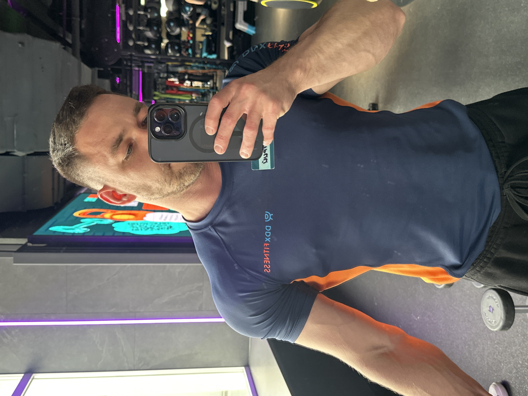 A man is taking a selfie in a gym, holding a phone with a dark grey case. He is wearing a dark blue and orange athletic shirt and black shorts. In the background is gym equipment. It’s likely the gym is named DDX Fitness. There is no other information to establish the exact location in Perm, Russia.