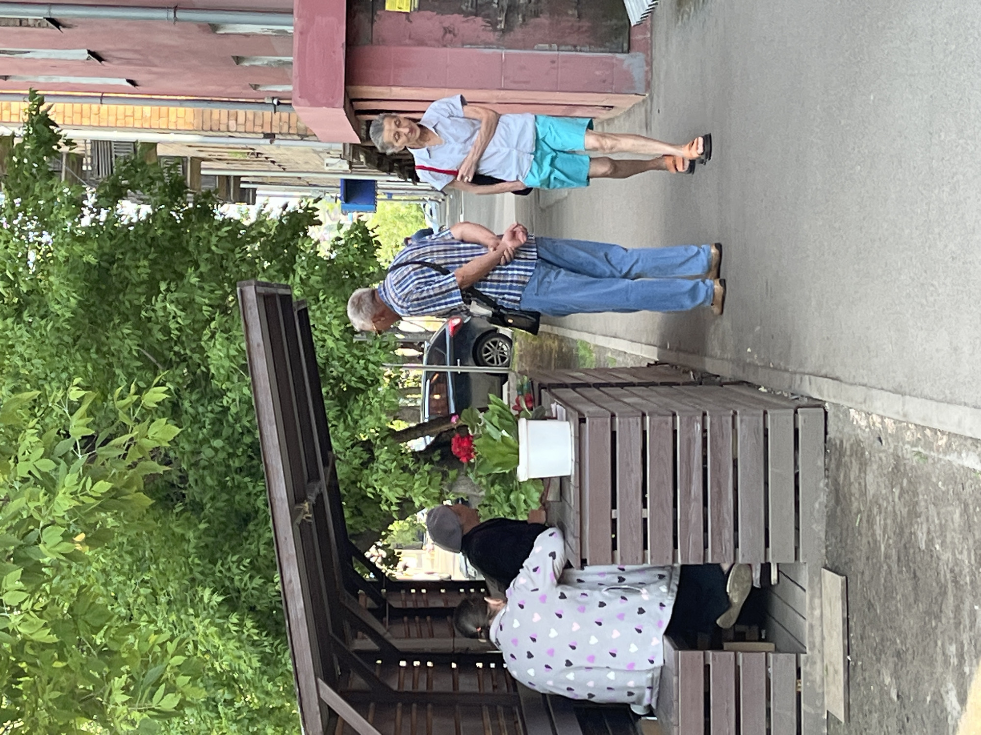 The image depicts a street scene in Krasnoyarsk, Russia, with an emphasis on outdoor seating and pedestrian activity. In the foreground, a wooden structure with a roof provides shade for two individuals seated on a bench. One person, viewed from the back, wears a light-colored jacket adorned with a pattern of pink and grey hearts. Their legs are not fully visible. The other individual, also facing away, is dressed in dark clothing and a grey cap. A white pot containing red flowers and green foliage sits on a wooden surface between them.
Further down the sidewalk, two more individuals are walking. A man in a blue and white plaid shirt and blue jeans stands facing the seated individuals, his hands clasped behind his back. A woman, approaching the viewer, is dressed in a light blue short-sleeved shirt and turquoise shorts. She carries a dark bag over her right shoulder and wears orange sandals.
The background reveals elements of an urban environment, including a tree with lush green foliage, a building with a pink facade, and a portion of a car. The street appears to be paved, with a wet surface in some areas, suggesting recent rain. The overall impression is of a casual, everyday moment captured on a street in Krasnoyarsk.