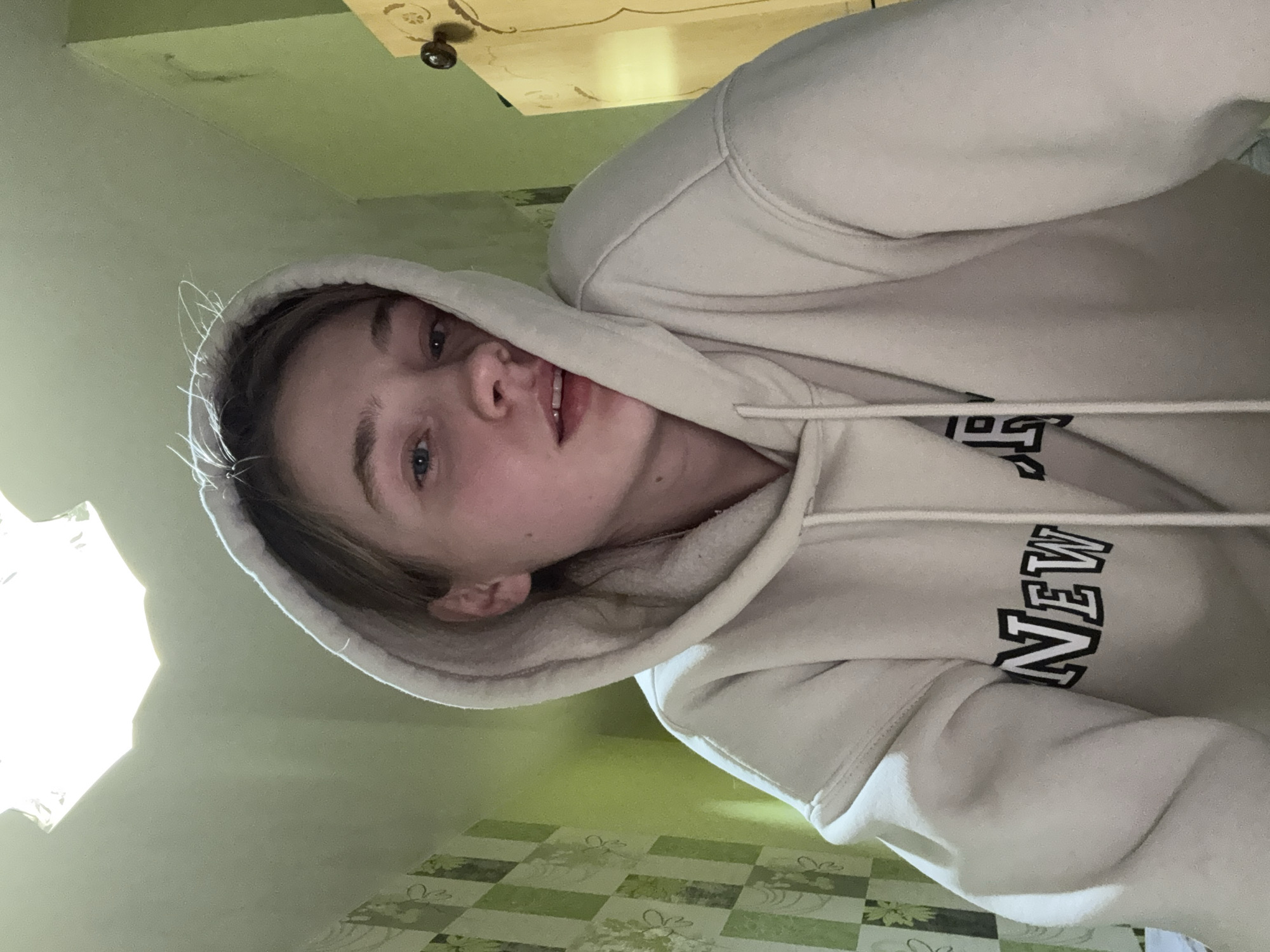 A young woman is wearing a beige hoodie with "NEW" written on the front. She is looking at the camera. The room appears to be in a home. The location is not provided in the image and can not be ascertained.