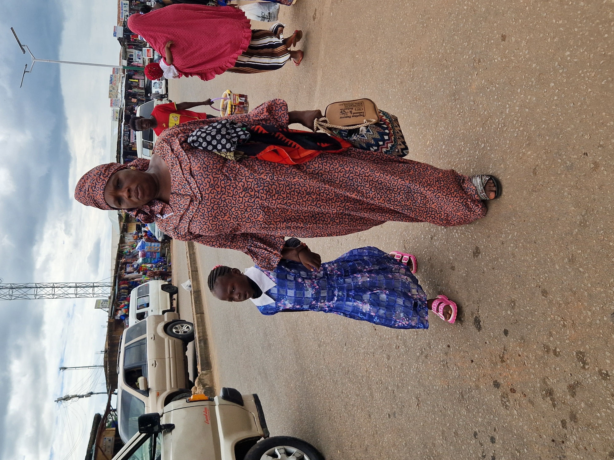 An eye-level, full shot captures a woman and a young girl walking on a paved street in Jos, Nigeria. The woman, in the foreground, wears a pink and orange patterned dress and a matching headscarf. She carries a brown purse and a patterned cloth bag on her left arm. To her left, the girl, dressed in a blue plaid dress with a white collar, holds the woman's hand. Her pink sandals are visible as she walks. Behind them, several vehicles are parked, including a gold SUV and a white van. The background reveals a marketplace with stalls lining the street, indicating a bustling urban environment. A large metal tower stands tall in the distance, and a cloudy sky completes the scene.