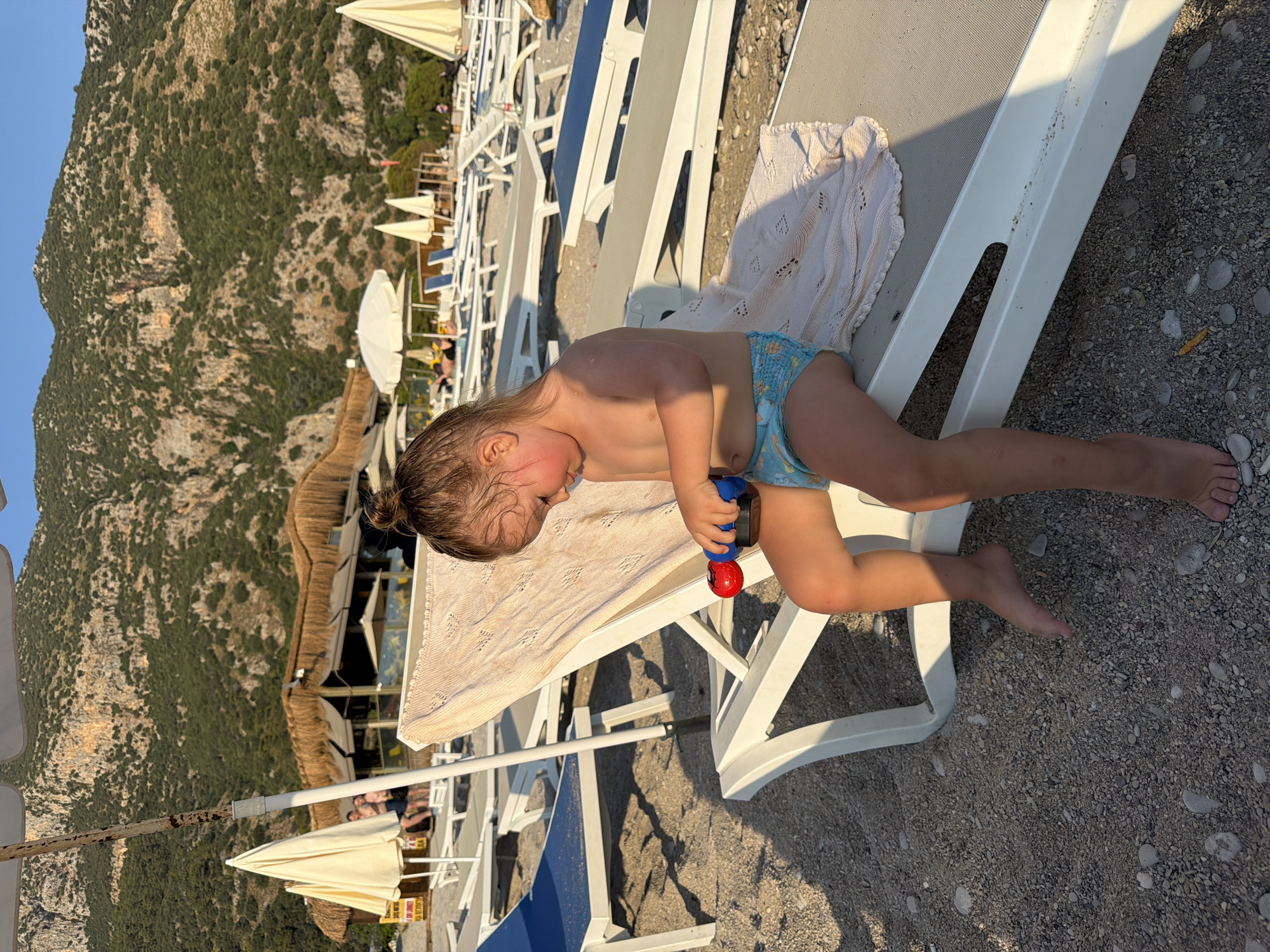 In Fethiye, Turkey, a young child sits on a white lounge chair on a pebbly beach. The child, facing left, is wearing a blue diaper with a printed pattern and is holding a red and blue toy. A light-colored, patterned throw is draped over the lounge chair. Behind the child, rows of empty lounge chairs and white beach umbrellas are visible, leading towards a thatched-roof structure with tables and chairs. In the background, a steep, tree-covered mountainside rises under a clear blue sky. The setting sun casts long shadows across the beach.